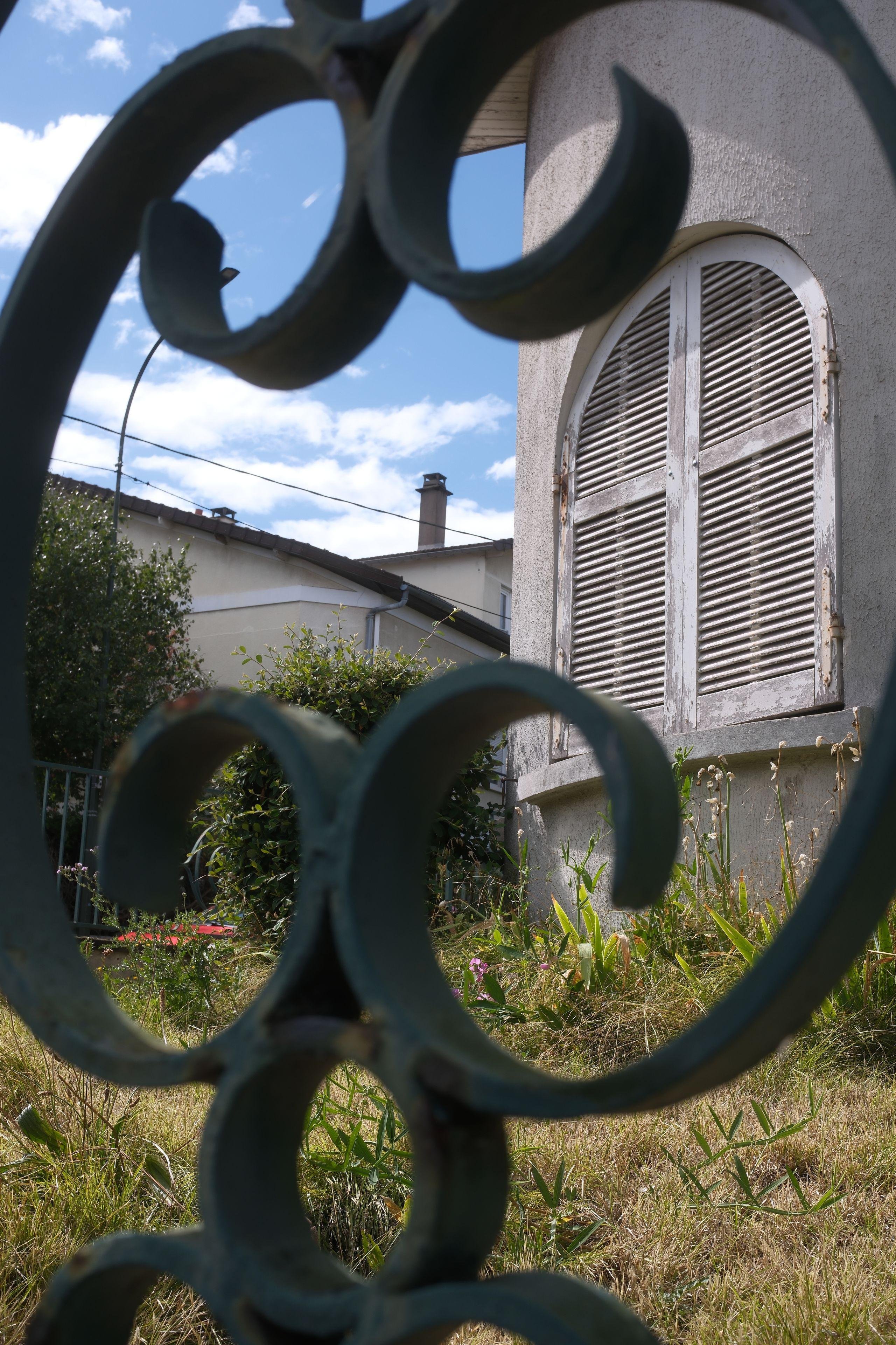 Photo format portrait, montrant à travers une clôture en fer forgé décoratif, un jardin avec de l'herbe haute et des fleurs, une maison avec des volets en bois fermés qui auraient besoin d’un coup de peinture, sous un ciel bleu partiellement nuageux.