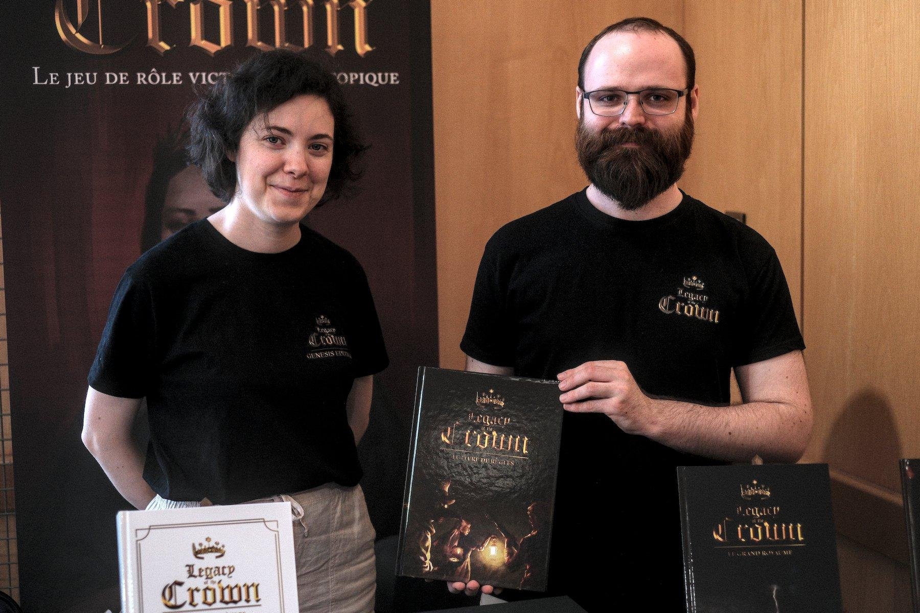 Photo horizontale. Portrait de face de deux personnes. À gauche une jeune femee aux cheveux noirs, à droite un homme barbu châtain tenant le livre de base de leur jeux de rôle, Legacy of the Crown