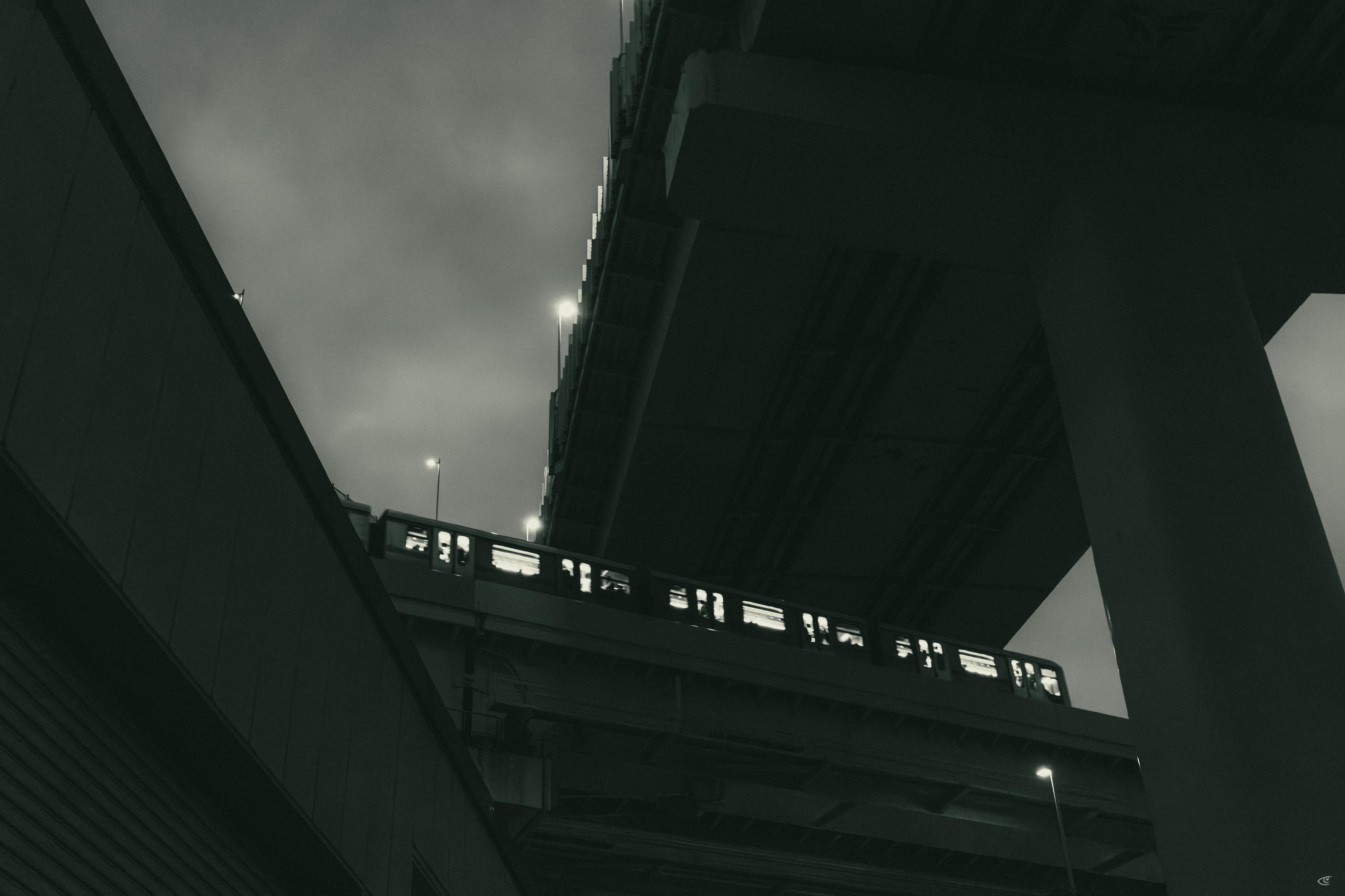 Low-angle view between two elevated overpasses, with a commuter train crossing a bridge section above; tall support columns frame the right side under an overcast sky.