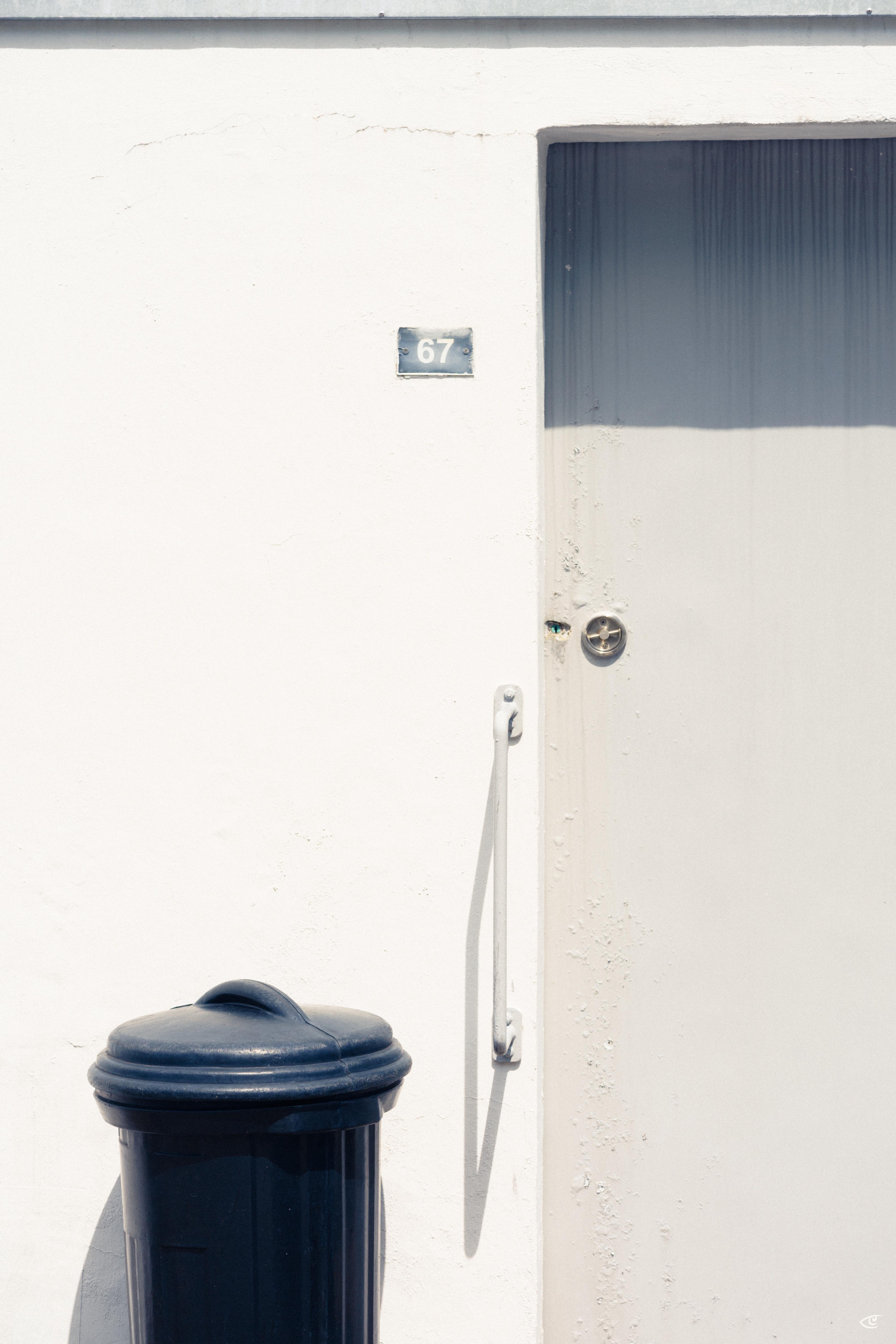 White exterior wall with a partially visible white door on the right, featuring a round metal knob and vertical handle; small plaque reading “67” above, and a dark blue trash bin in the lower left casting a shadow.