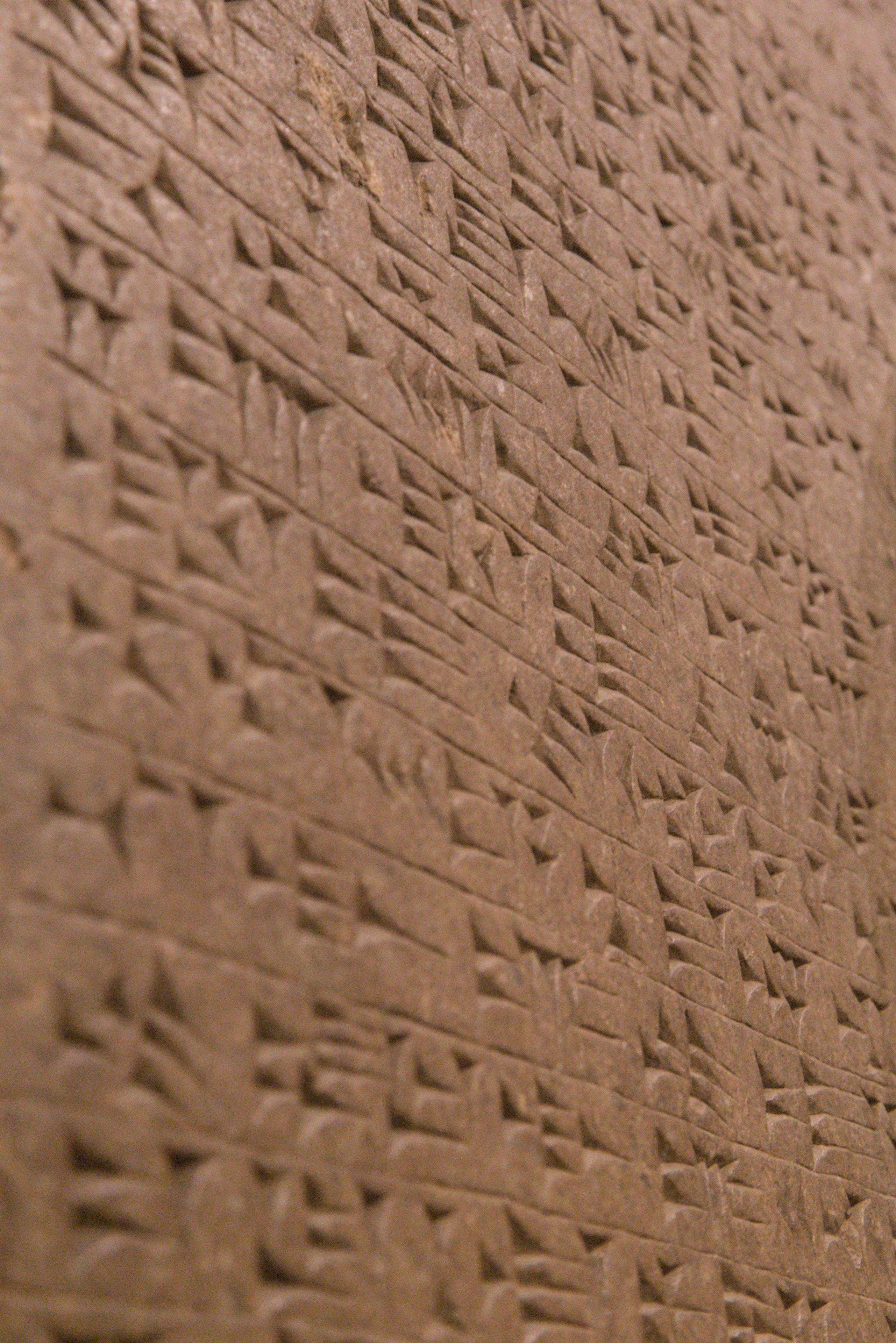 Photo format portrait. Gros plan de biais sur un mur de couleur marron avec des écrits cunéiformes. La profondeur de champs est fine faisant que seule une petite partie est nette, avec le reste en bokeh