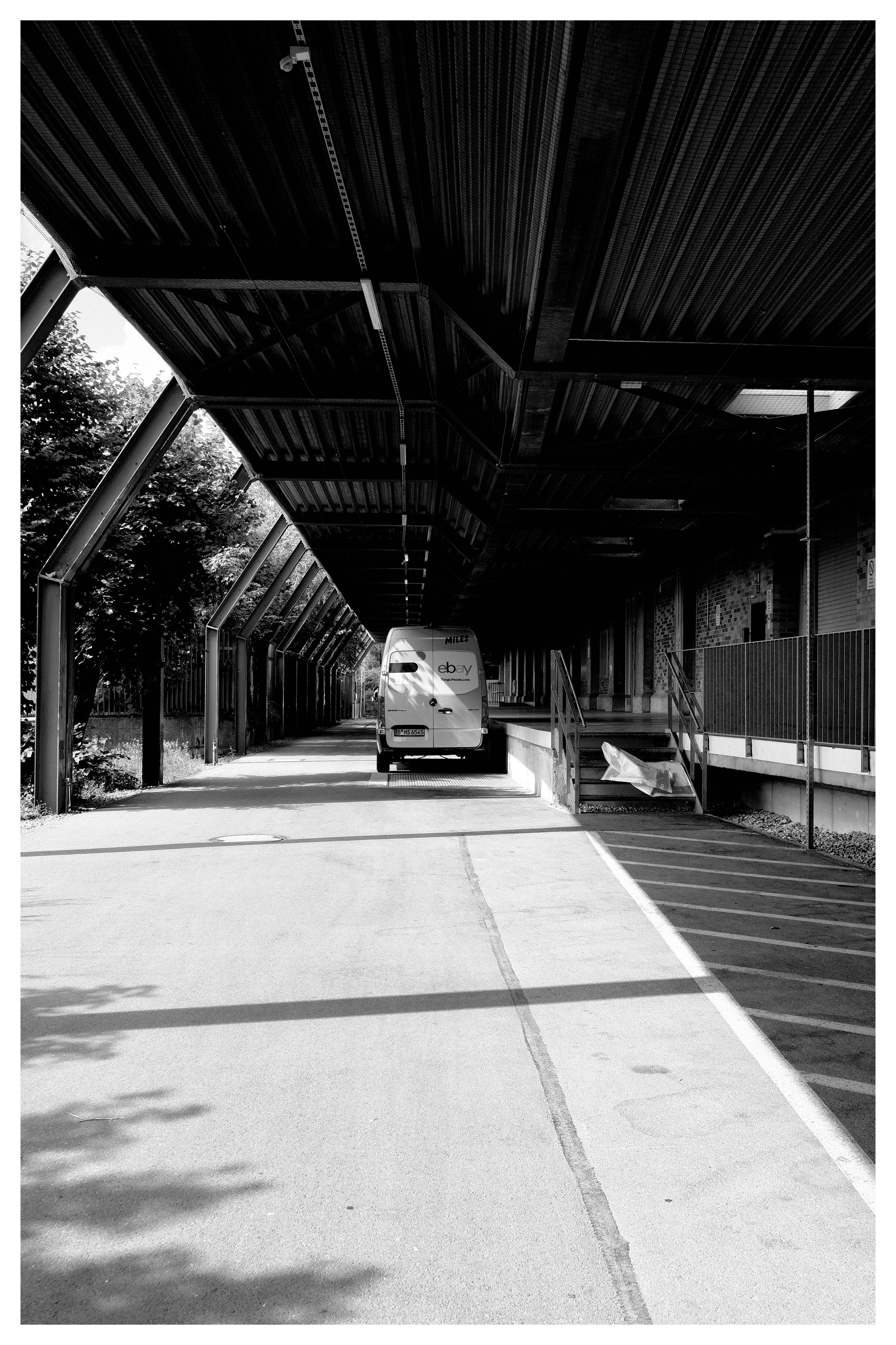 A black and white image of a van parked under a long, covered loading area. The structure has metal beams and a corrugated roof. Trees and foliage are visible on the left, while the right side shows a raised platform with a metal railing and a ramp. Shadows are cast on the ground, indicating sunlight from the left side.