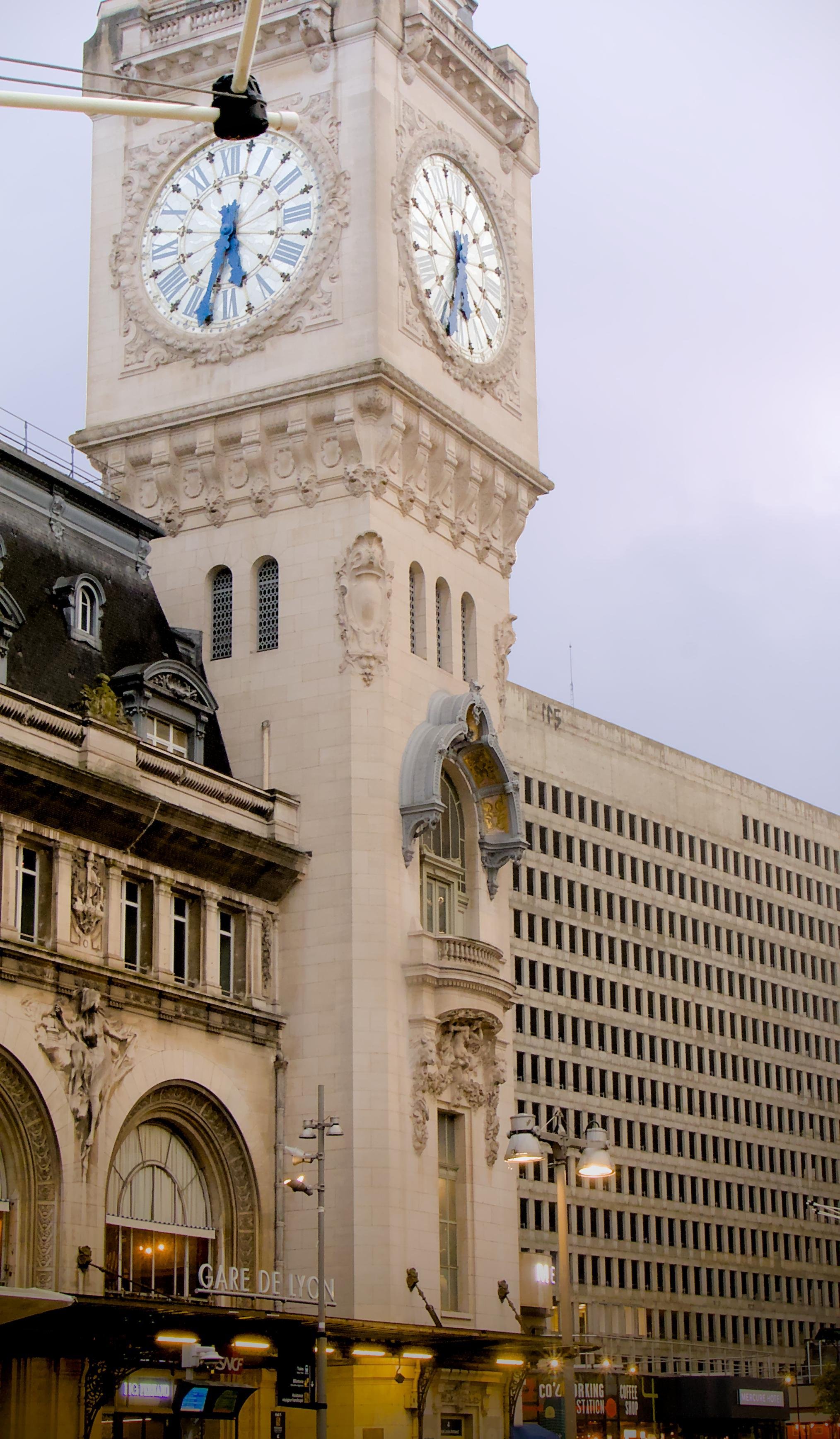 La photographie représente la tour de l'horloge de la gare de Lyon à Paris. La tour présente une architecture ornée de cadrans d'horloge sur plusieurs côtés. Le bâtiment de la gare est visible dans la partie inférieure de l'image, y compris une partie de sa façade détaillée et de ses éléments décoratifs. Un autre bâtiment est visible sur la droite de l'image, avec une architecture neutre, un bloc de béton avec plein de fenêtre. La différence d'architecture crée un contraste dans l'image.