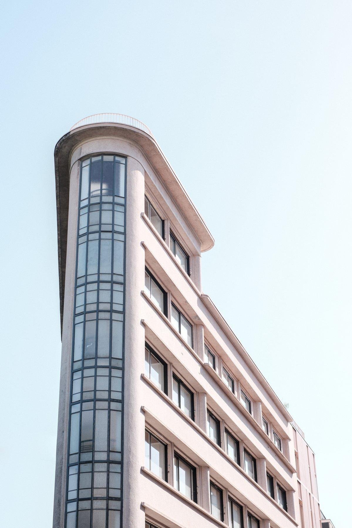 Photo verticale. Un immeuble rose pale à l’architecture sortant de l’ordinaire, étroit à un bout et qui s’évase en triangle, sûrement pour suivre la chaussée qui est hors cadre. La pointe du bâtiment est arrondie et avec du verre/acier noir tout le long. Le tout sur fond de ciel bleu pastel