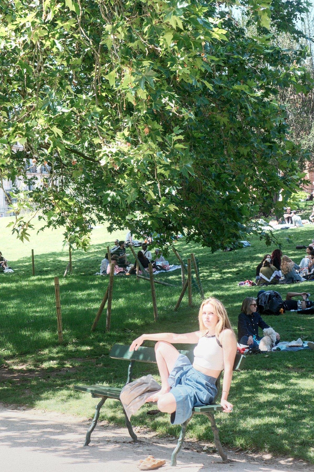 Photo verticale. Sur un banc au soleil dans un parc, une jeune femme blonde aux cheveux long est installée relax, une jambe en tailleur, l’autre le genoux en l’air, son bras droit posé dessus, le bras gauche posé sur le banc pour se tenir droite. Elle a remarqué la caméra. Derrière, la pelouse ombragée avec des groupes de gens assis dessus.