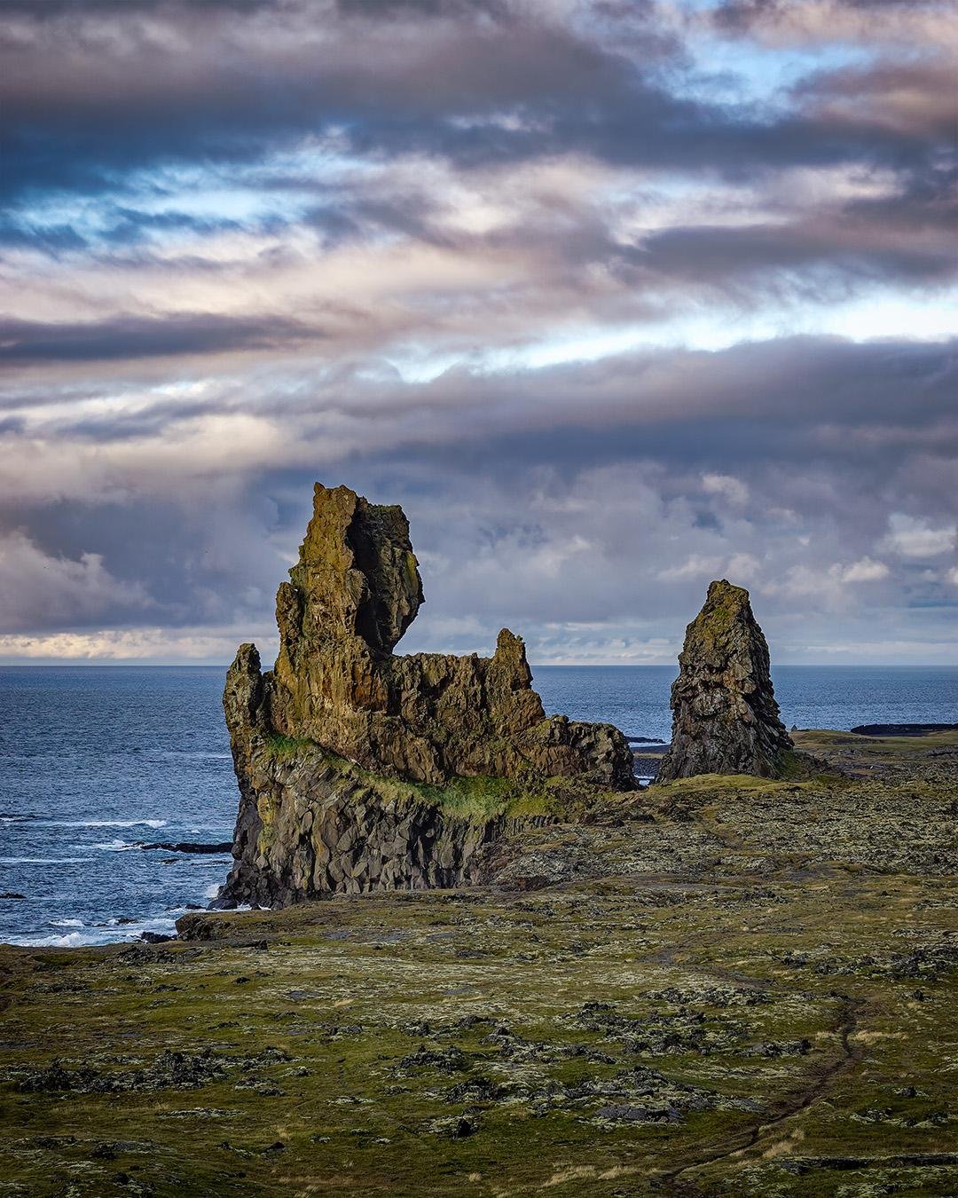 Rocky coastline with two prominent jagged rock formations rising from the sea. The foreground features a grassy, rugged terrain. The sky above is filled with dramatic clouds, with patches of blue visible, creating a contrast with the dark, textured rocks. The ocean in the background stretches to the horizon.