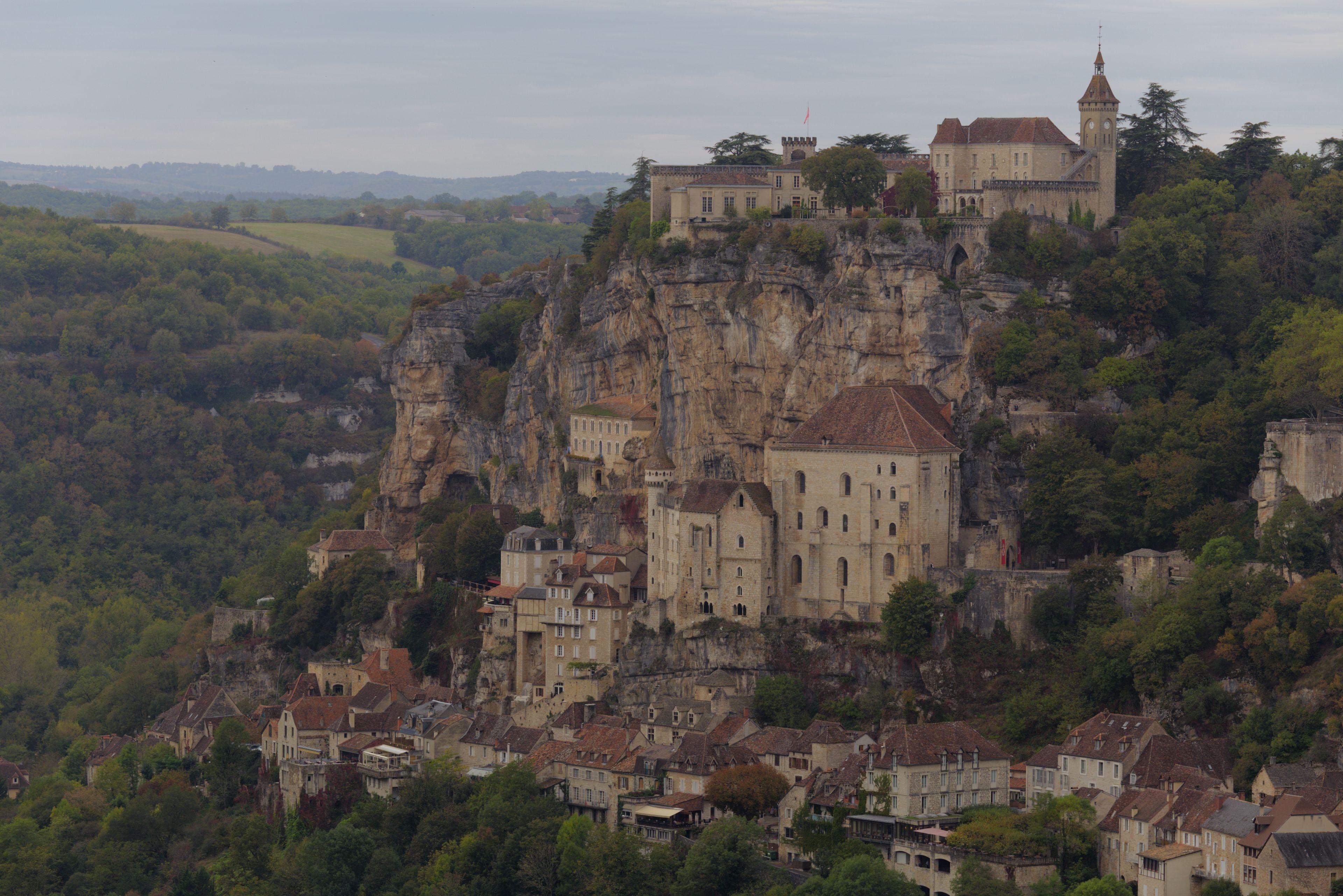 Photo format paysage montrant le village de Rocamadour, construite au pied mais aussi sur les falaises du coin. Avec la forêt en contre bas et au fond de l’image