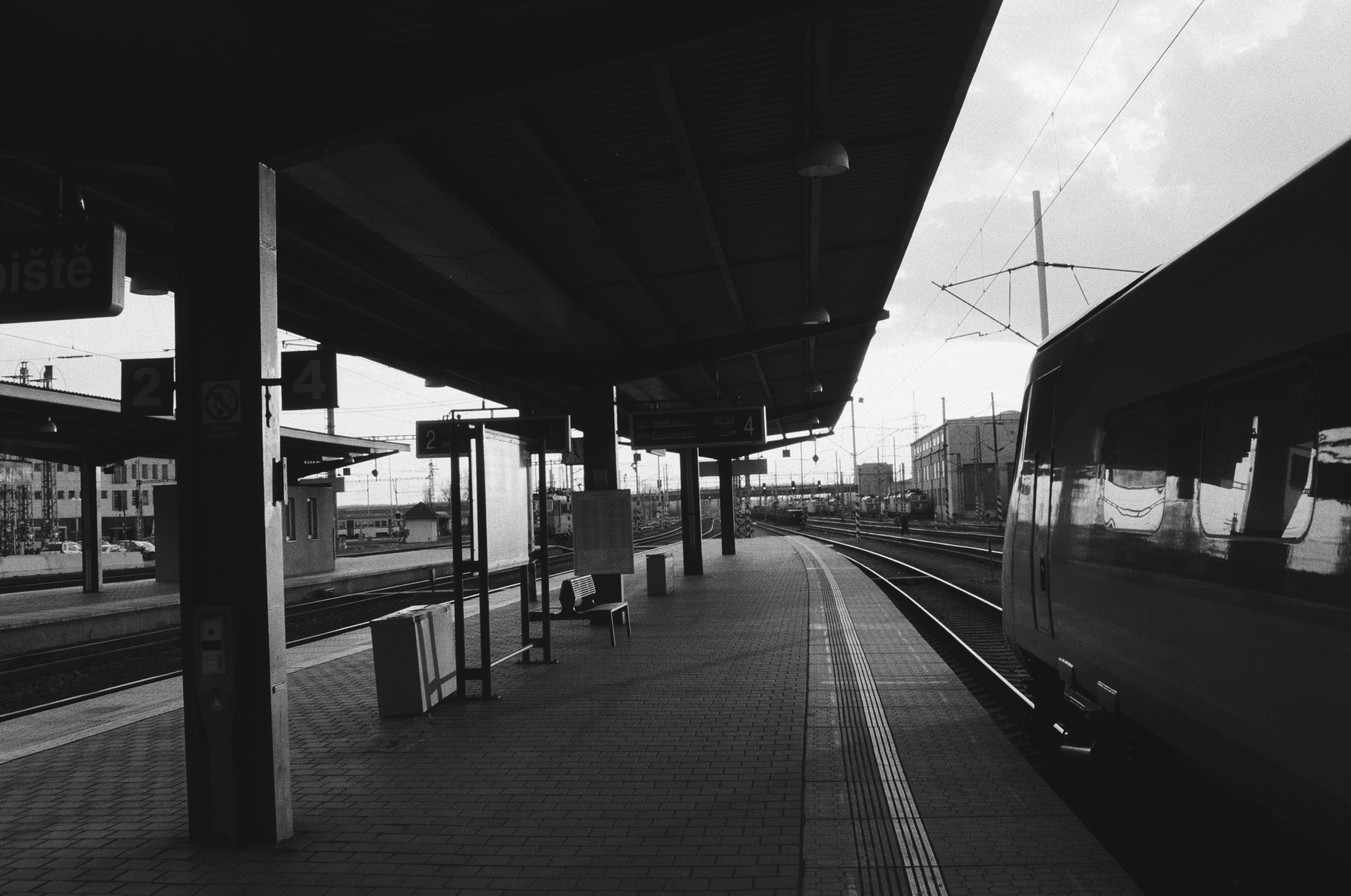 A monochrome photo of a train platform with a dark roof and a train on the left side. There is a striking contrast between the dark of the train and platform and the lightness of the sky.