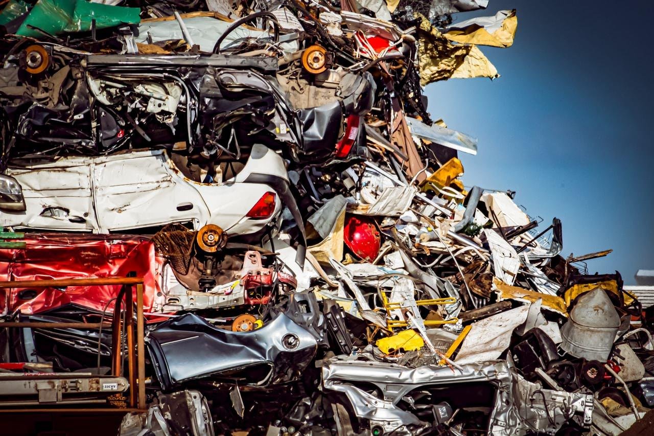 The image shows a large pile of crushed and dismantled cars, likely at a scrapyard. Various car parts, such as metal panels, tires, and bumpers, are visible in the heap. The scene suggests recycling or disposal of vehicle materials.