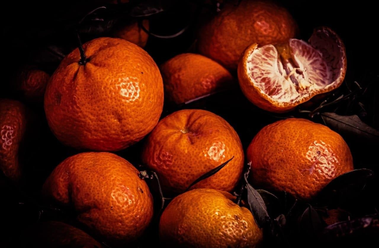 Cluster of whole mandarins with dark green leaves on a dark background, with one mandarin partially peeled open at the top right showing pale segmented flesh.