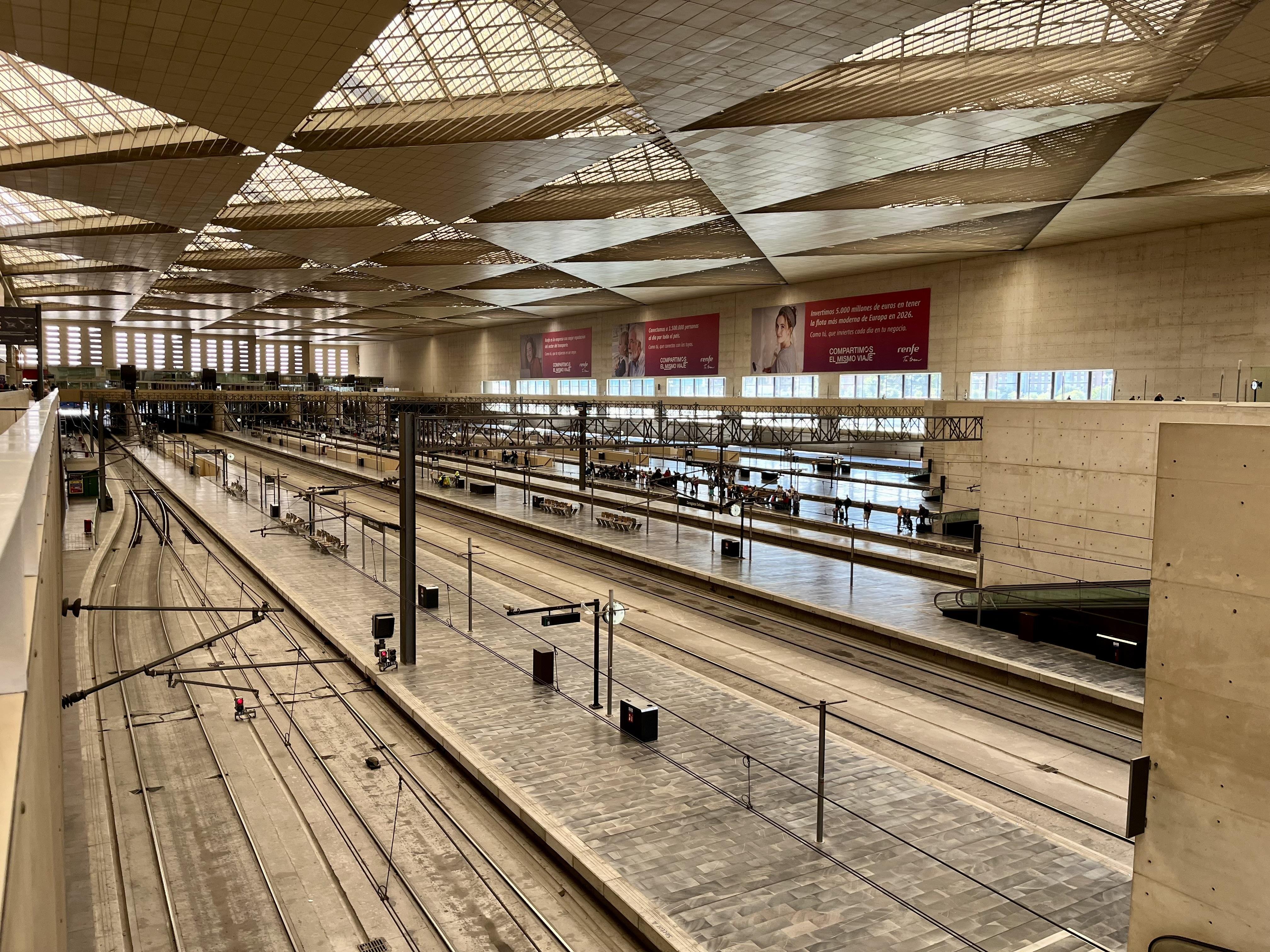 This image shows an empty railway station with multiple tracks and platforms. The architecture of the station features a high ceiling with large windows allowing natural light to enter. There are signs and advertisements on the walls, and the overall space appears spacious and modern.