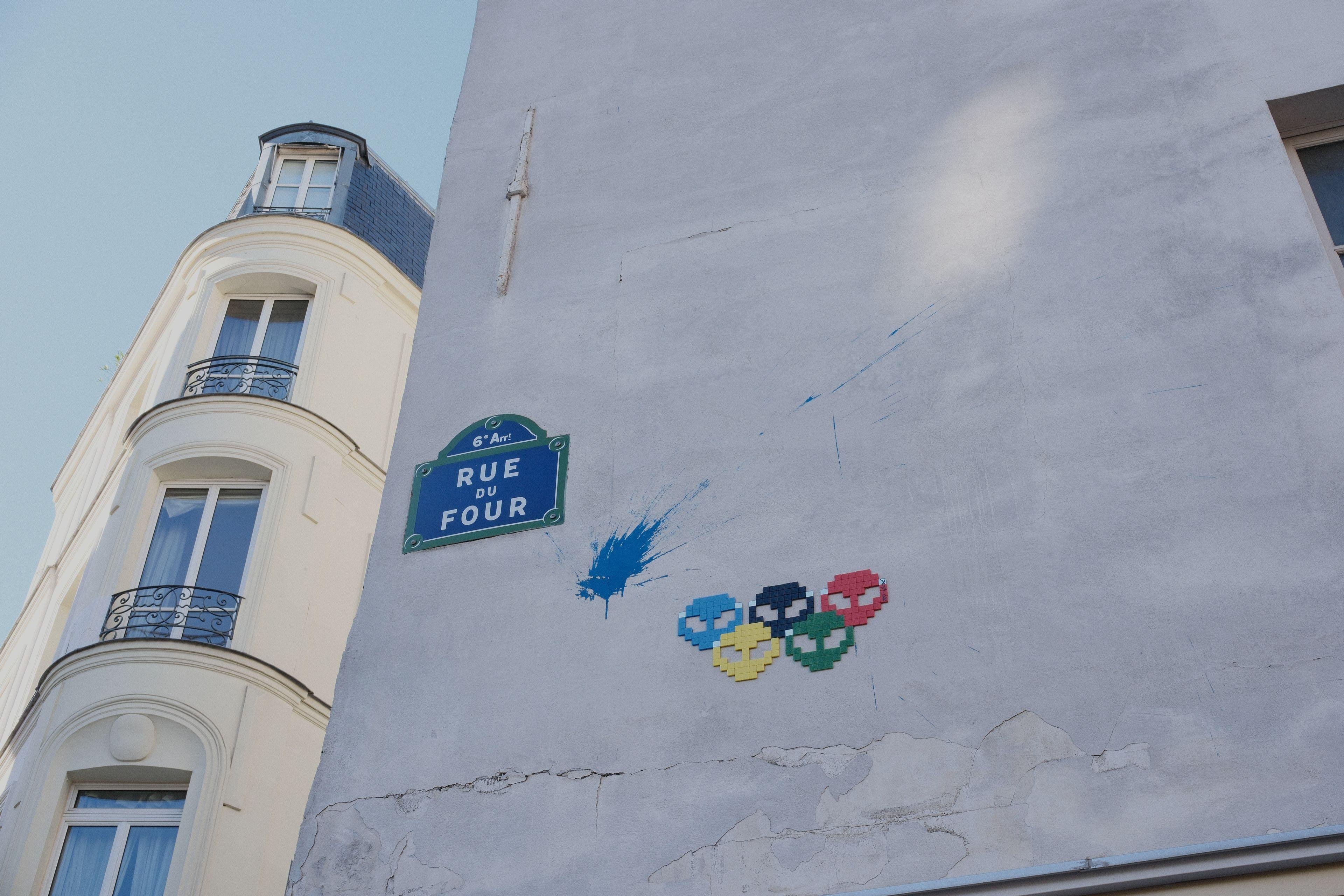 Une mosaïque formée de 5 têtes d’alien disposées comme des anneaux olympiques de couleurs bleu, jaune, noir, vert, rouge. À gauche, un splash de peinture bleue, et encore à gauche, une plaque de nom de rue « Rue du Four », puis à gauche, le mur laisse place à un immeuble parisien ensoleillé