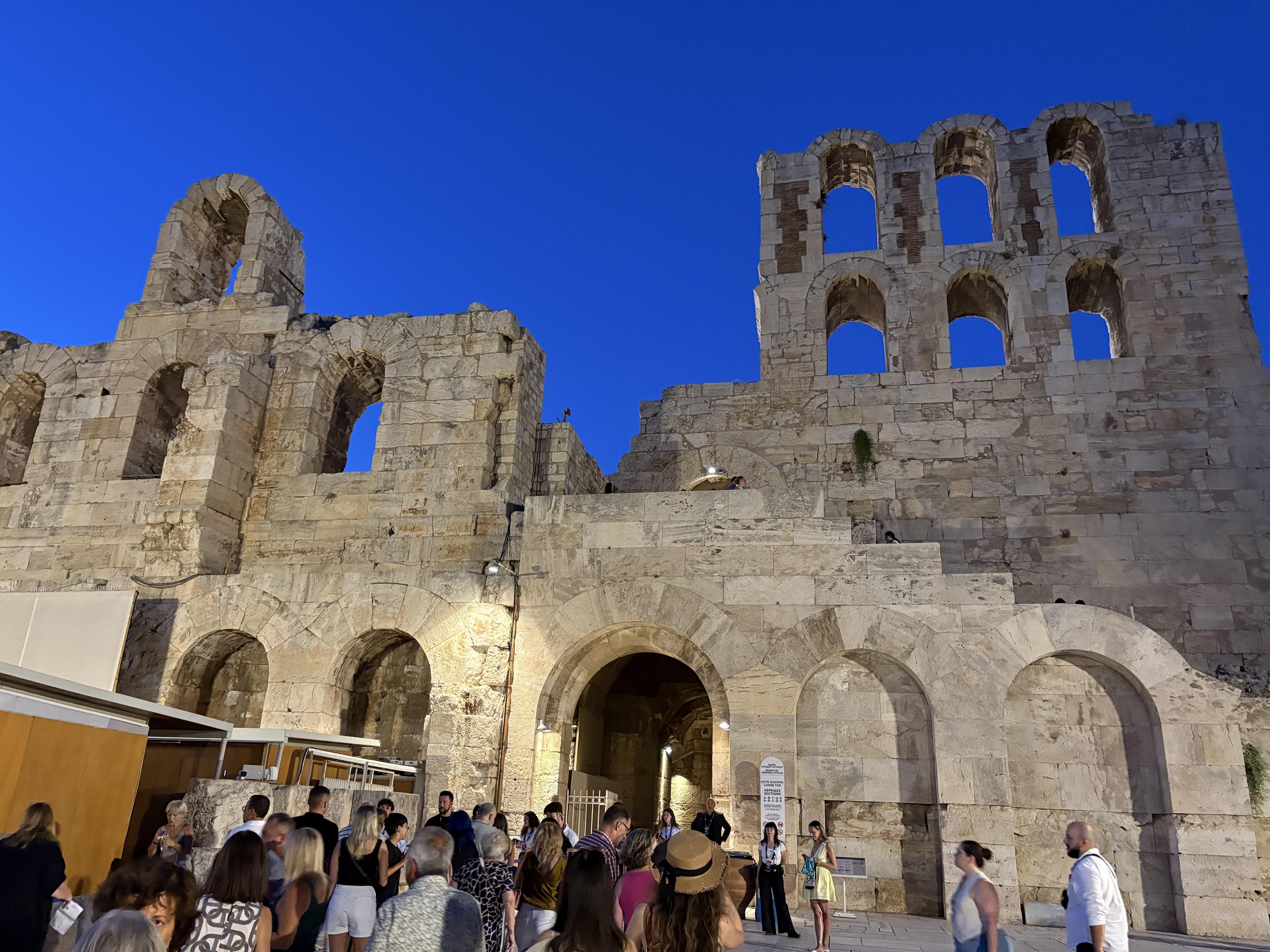 Ancient stone structure with multiple arched windows and doorways illuminated against a vivid blue sky. A large group of people gathers in front, indicating a lively atmosphere. The stone walls show signs of aging, adding to the historical ambiance.