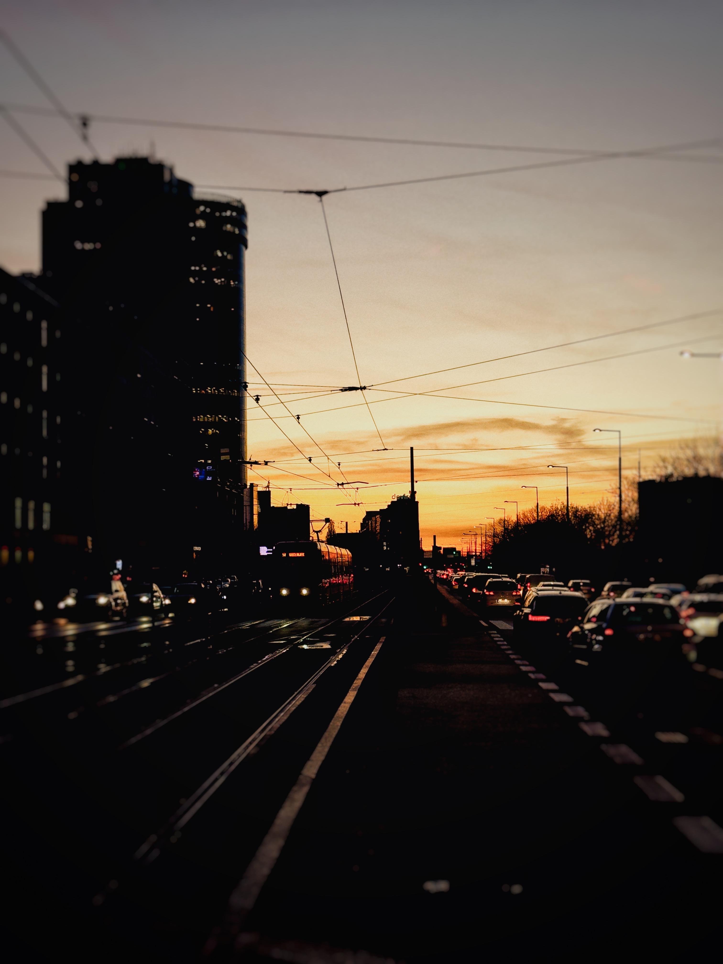 City landscape of Warszawa during sunset. On both sides there are cars staying in the traffic, in the middle there is a tram at the tram stop. One tall building visible in the background. The sky is orange–ish at the bottom and yellow–ish at the top of the picture.