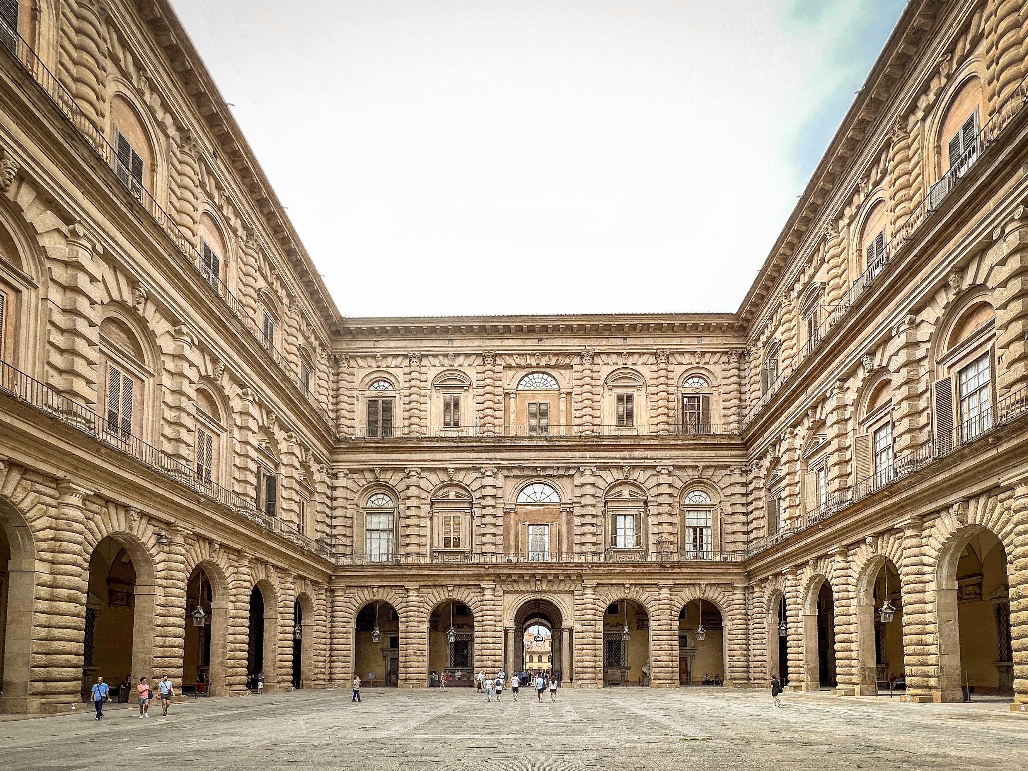 Corte interna di Palazzo Pitti a Firenze, con la sua imponente architettura rinascimentale. La corte è ampia e lastricata, con facciate in pietra che si sviluppano su più livelli, caratterizzate da archi e finestre. Si possono vedere persone che passeggiano nella corte, dando un senso di scala all'edificio. La luce del cielo illumina l'intera scena, mettendo in risalto i dettagli architettonici del palazzo.