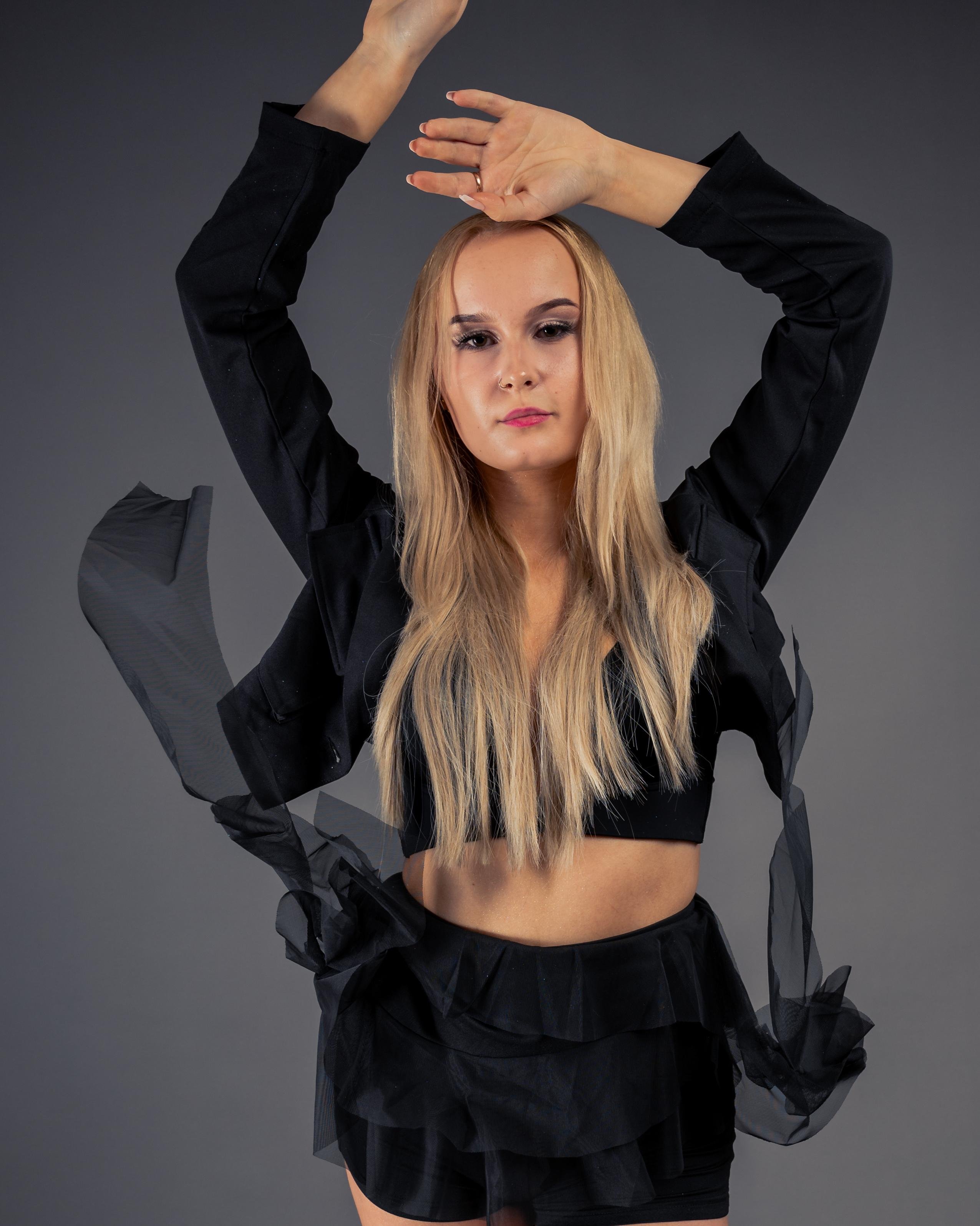 Dancer with long blonde hair wearing a black crop top and layered skirt, raising arms gracefully against a gray background. Translucent fabric accents the outfit, creating a fluid appearance.