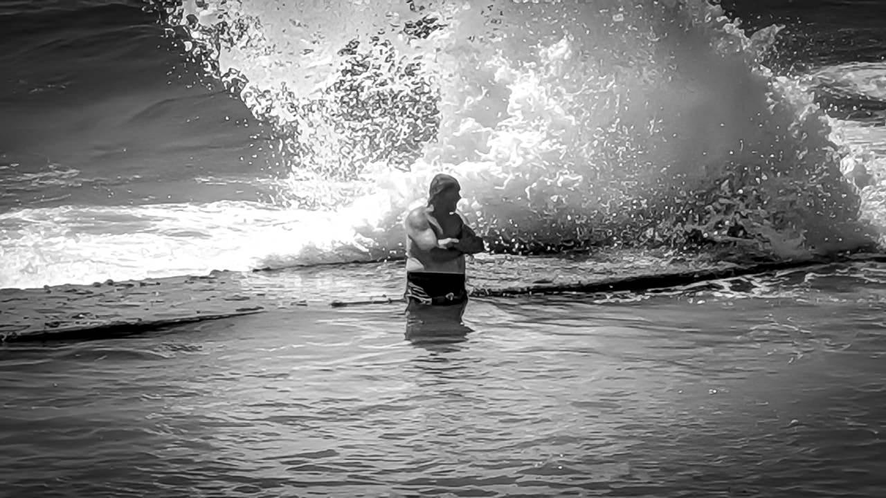 A man stands waist-deep in water near the edge of a large wave crashing against rocks. He is shirtless with his arms crossed over his chest. The image is in black and white, highlighting the contrast between the dark water, the man's figure, and the white froth of the wave.
