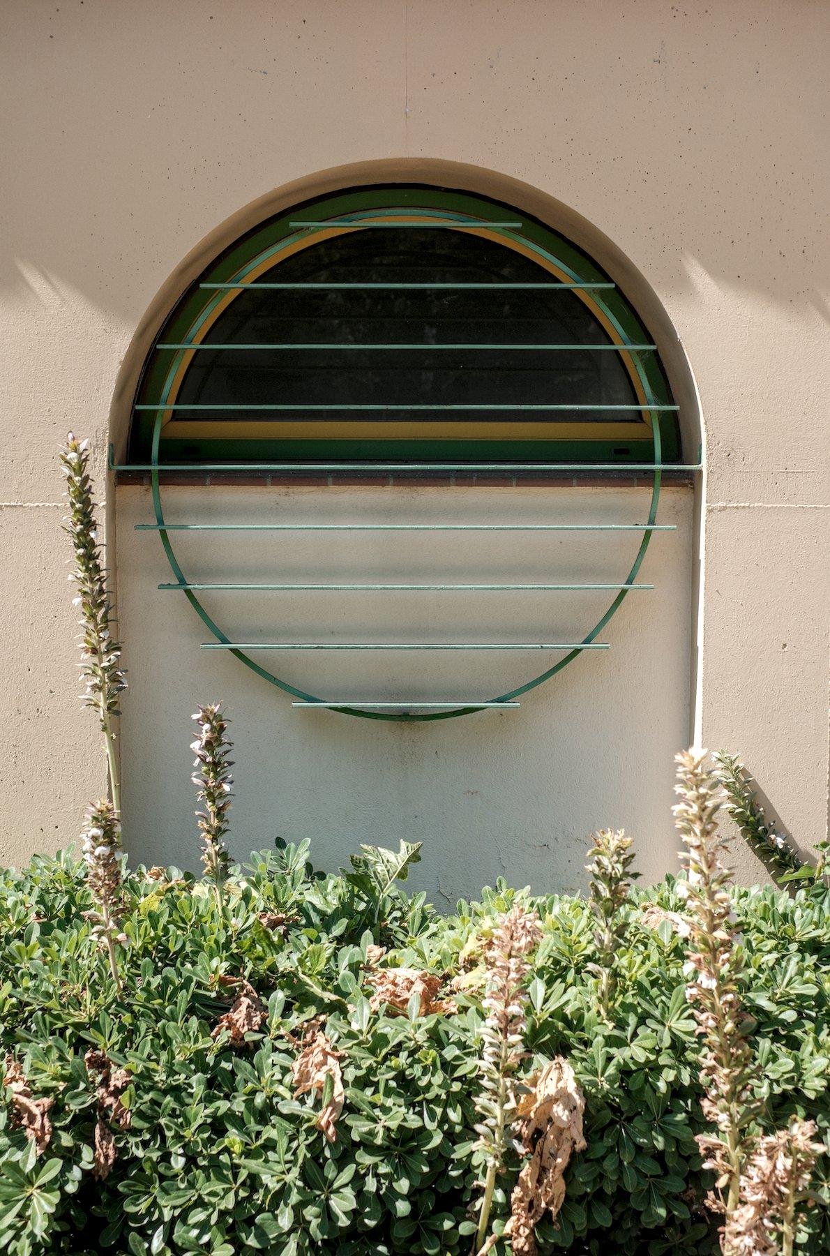 Photo verticale. Derrière un parterre de végétation, dans un mur beige est creusé une fenêtre au sommet arrondi, avec une grille verte circulaire aux barreaux horizontaux, protégant une fenêtre ne faisant que la moitié supérieure du cercle