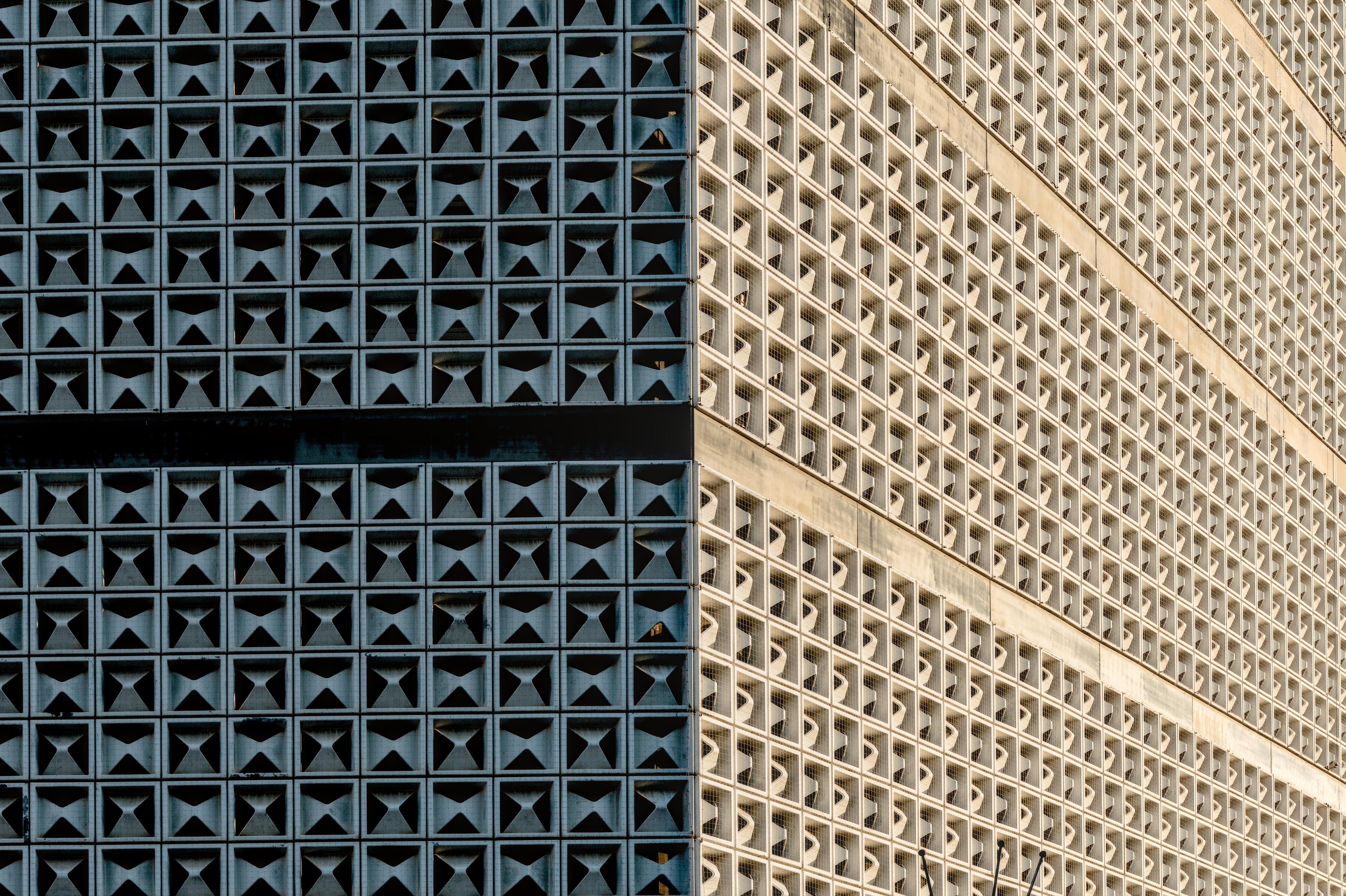 Two adjoining building facades in close-up, divided by a vertical seam: left side dark blue with a grid of square recessed openings, right side light beige with a similar square lattice extending diagonally into the distance. ###
Zwei angrenzende Gebäudefassaden in Naht, geteilt durch eine vertikale Naht: linke Seite dunkelblau mit einem Gitter aus quadratischen Einbauöffnungen, rechtsseitiges Lichtbeige mit einem ähnlichen quadratischen Gitter, das sich diagonal in die Entfernung erstreckt.