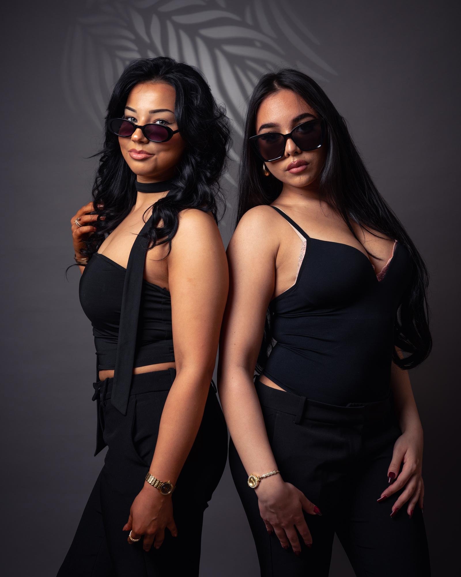 The image shows two individuals standing back-to-back. They are both wearing black outfits and sunglasses. The background is a neutral gray with a leaf pattern shadow. They have long hair and are posing confidently. Valokuvaaja Kuvaajankulma