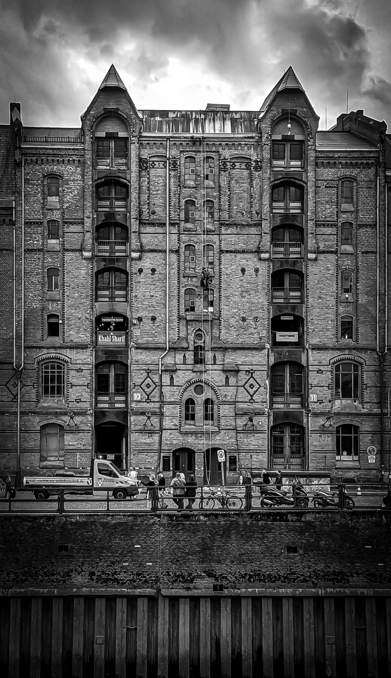 Black-and-white, front-facing view of a tall brick warehouse with arched windows and two pointed corner towers under a cloudy sky; a street with pedestrians, bicycles, a van, and scooters runs along the base behind a railing and canal wall.