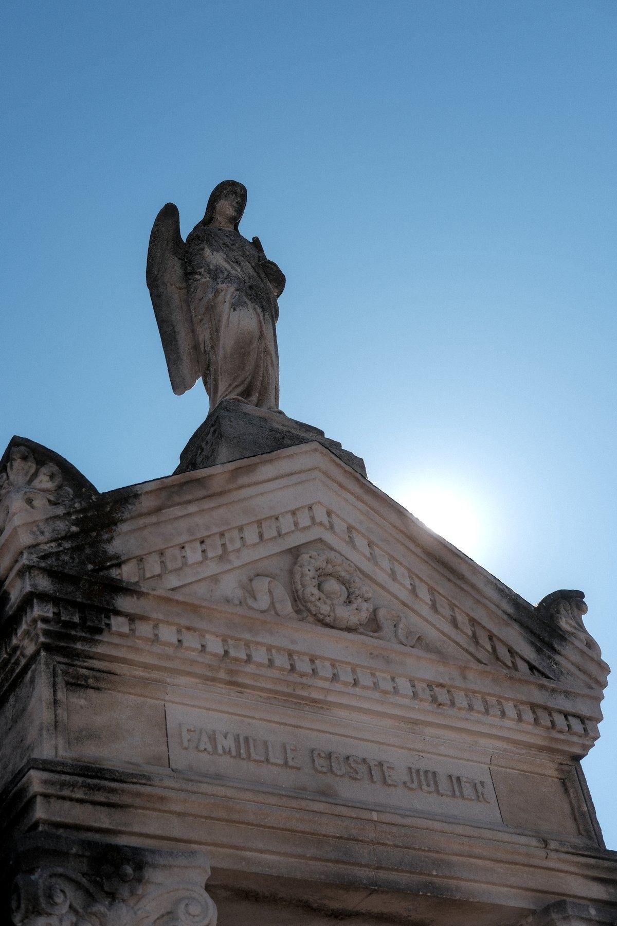 Photo verticale. Statue d’un ange au sommet d’un mausolée sur fond de ciel bleu. Le soleil dépasse légèrement «&nbsp;toit&nbsp;» du mausolée sur la droite
