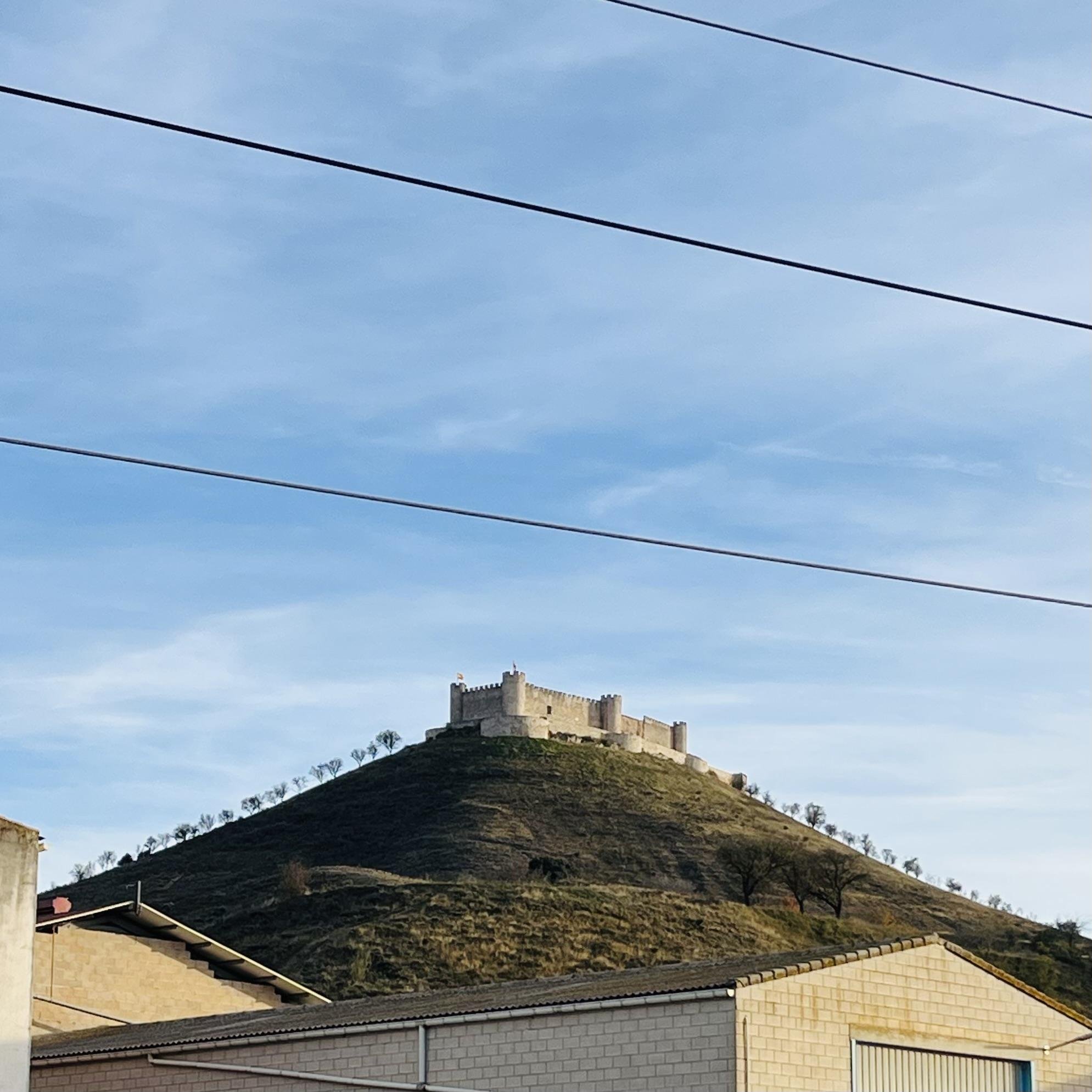 The image shows a hill with a castle on top, surrounded by greenery and trees. In the foreground, there are some buildings, and power lines cross the view. The sky is clear with few clouds.