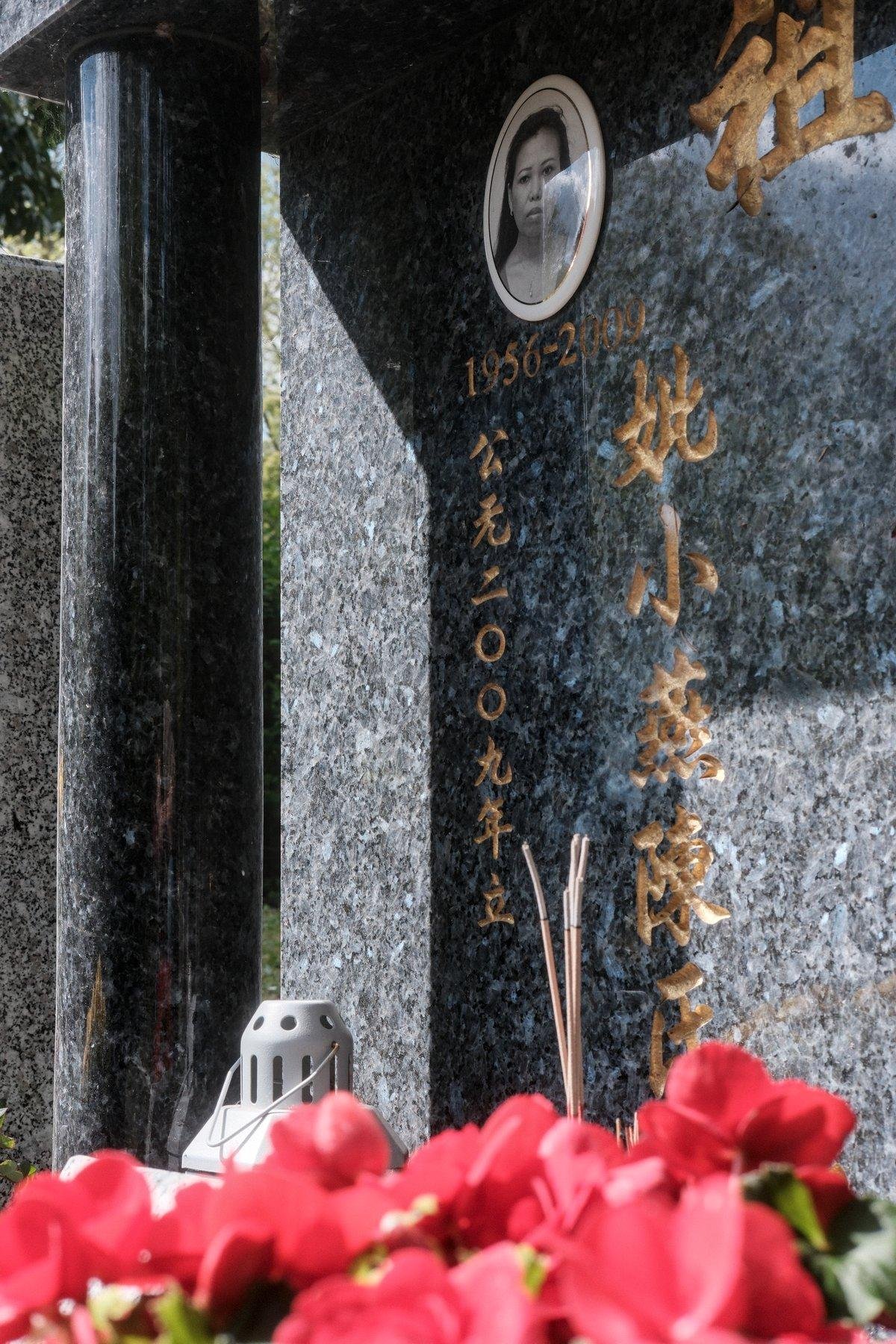 Photo verticale. Au premier plan, des fleurs rouges floues, derrière elles, une pierre tombale en marbre avec des écritures en chinois et le portrait de la défunte (ma mère). Des encens sont entrain de brûler sous le portrait