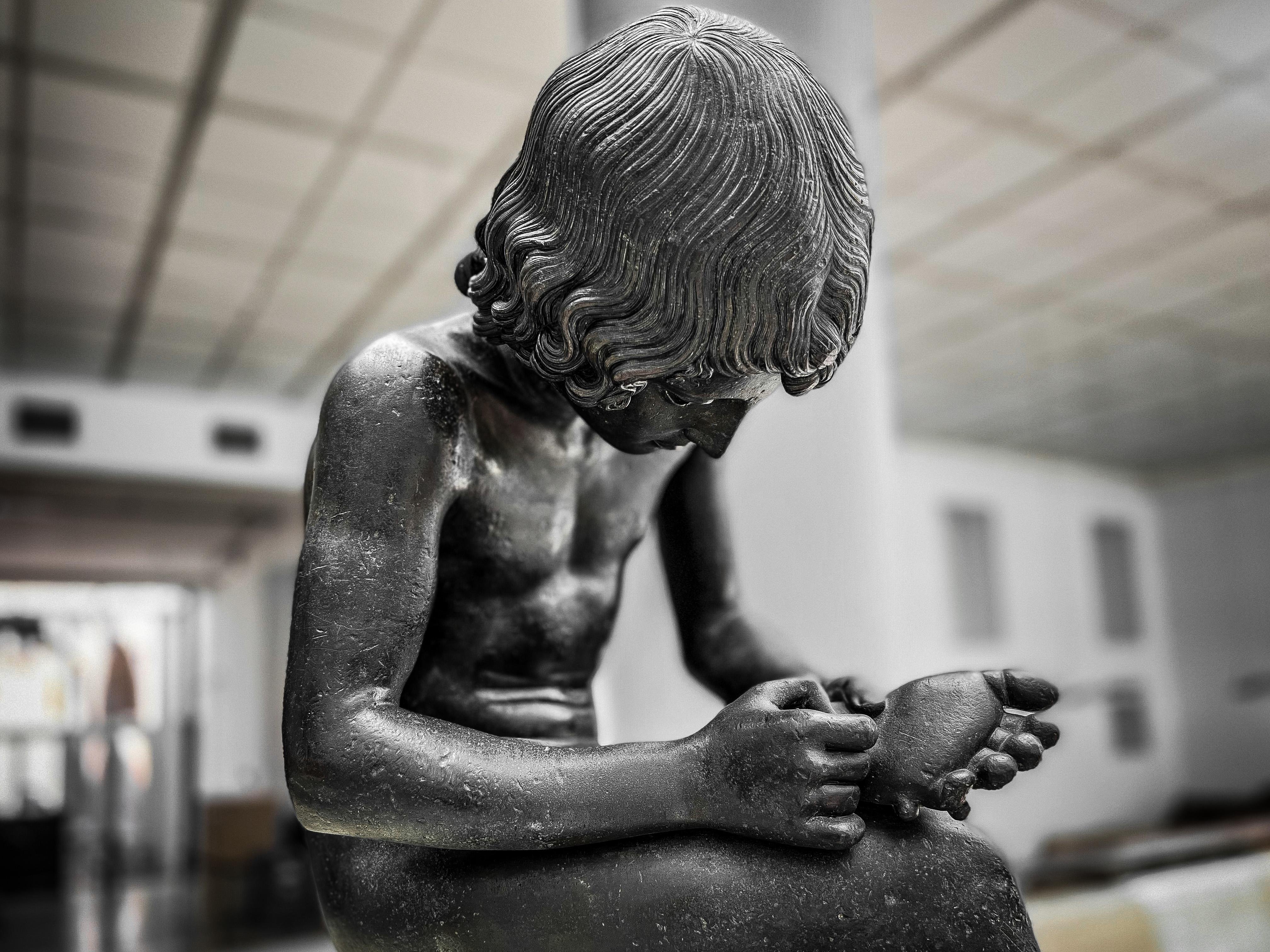 Scultura di un ragazzo seduto e con lo sguardo rivolto verso il basso. Il ragazzo sembra tenere in mano una spina, come si vede dal gesto delle sue mani. L'opera è conosciuta come "Il Spinario" o "Ragazzo con la spina," una famosa antica scultura che raffigura un ragazzo mentre rimuove una spina dal suo piede.