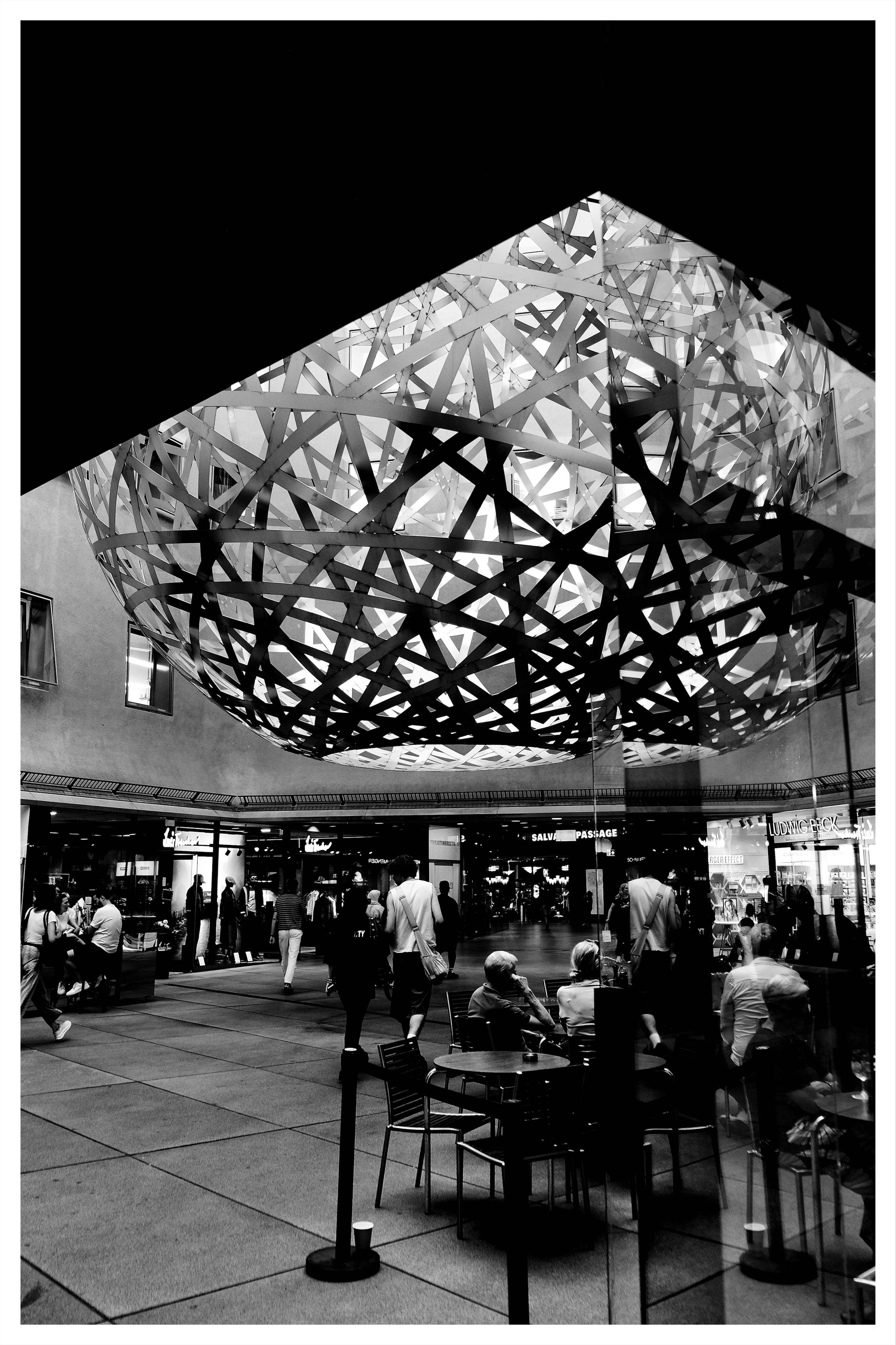 Black-and-white indoor plaza with a large illuminated spherical lattice sculpture suspended overhead; people walk and sit at café tables in the foreground, with shopfronts and a curved balcony lining the background.