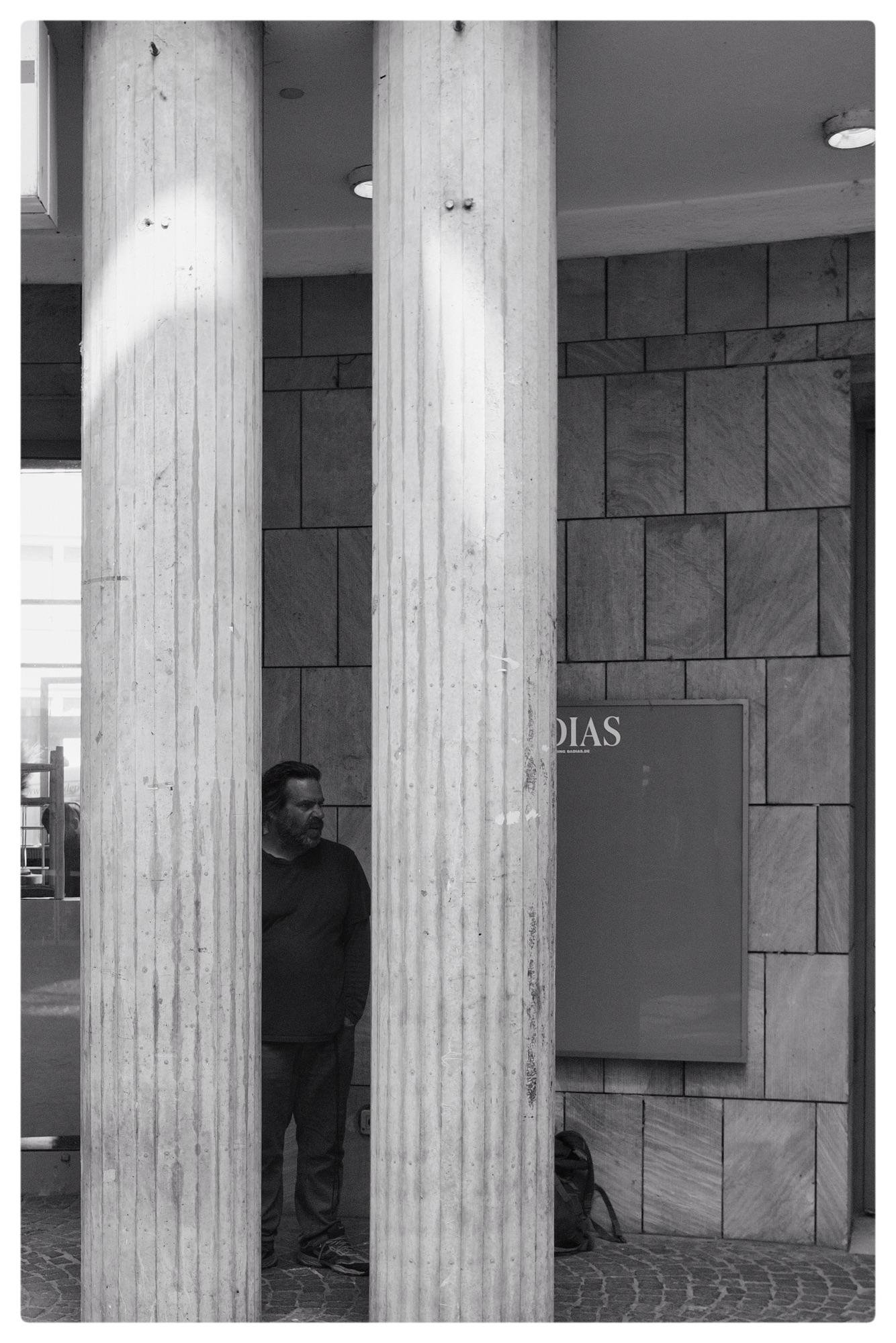 Black and white image of a man standing between two large stone columns with a textured surface. The man is partially hidden behind the columns and wearing a dark shirt and pants. The background features a wall with square tiles and a partially visible sign with text. A backpack is placed on the ground near the wall.