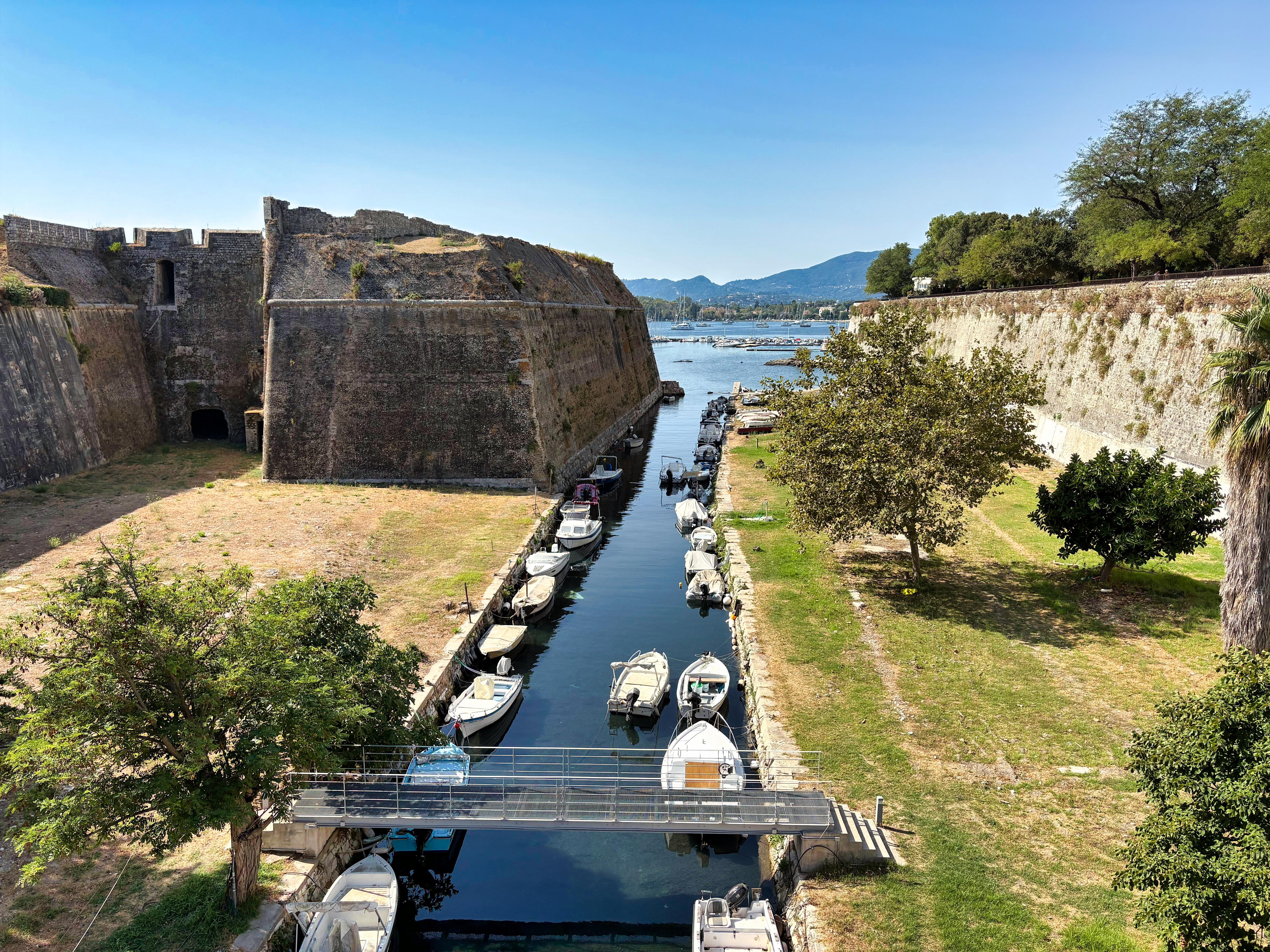 Narrow water canal lined with small boats flows between two ancient stone walls, leading towards a larger body of water. On the left, a large fortress structure with grassy terrain. On the right, a grassy area with trees. A small bridge crosses the canal. Hills and a clear blue sky are visible in the background.