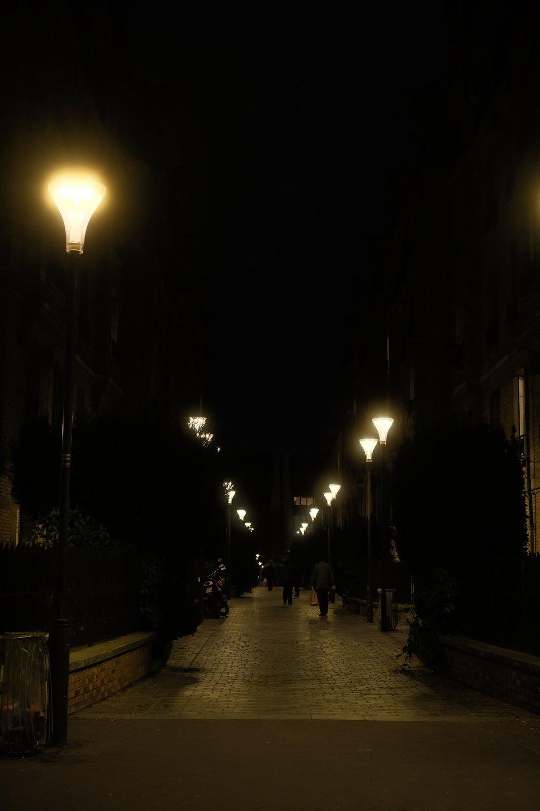 Photo format portrait, de nuit, d’une allée piétonne pavée, éclairée par des lampadaires, encadrée de petits immeubles, des gens de dos marchent dessus