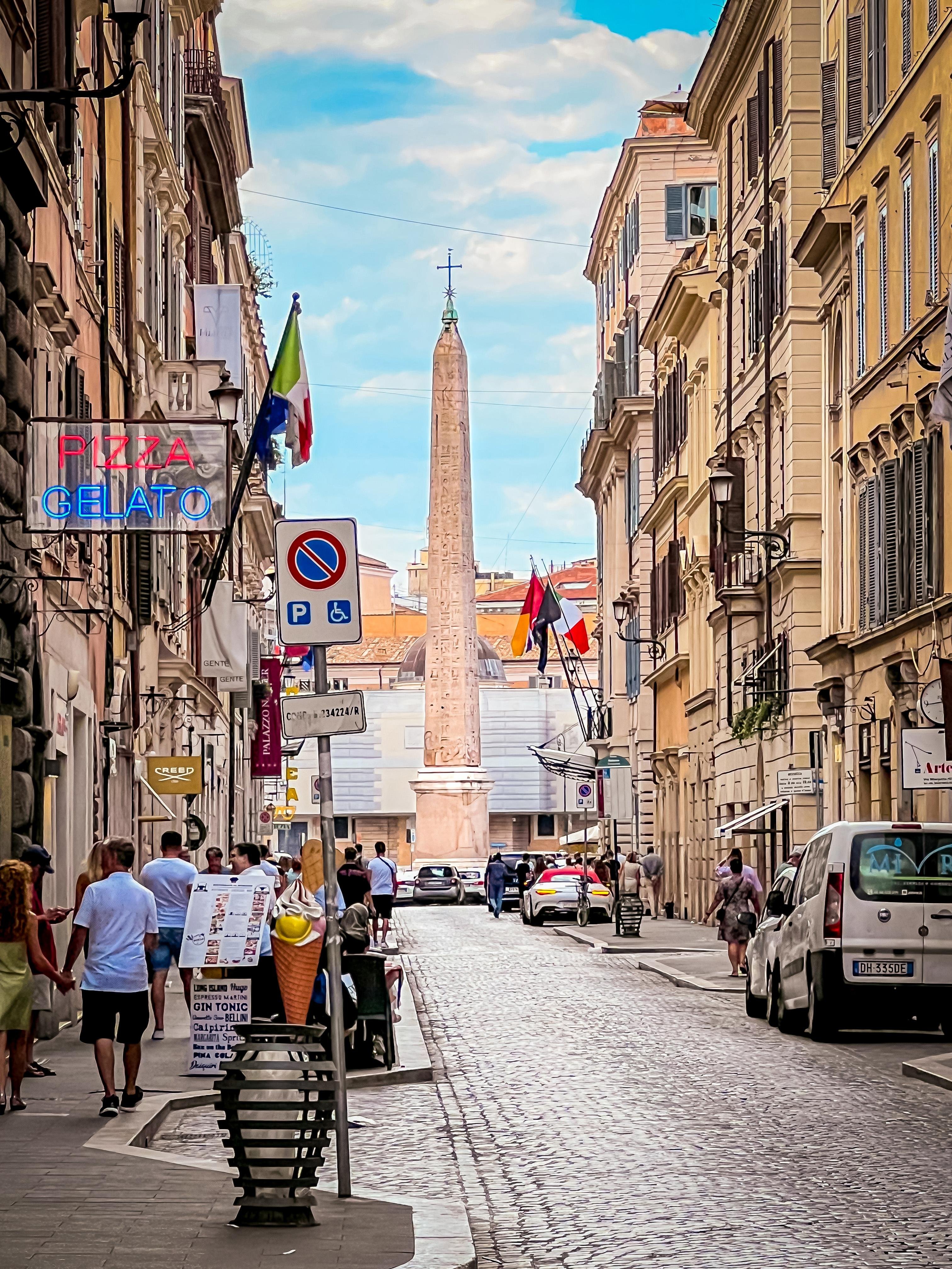 Strada stretta fiancheggiata da edifici, probabilmente in una città europea. Sono visibili bandiere italiane, suggerendo che si trova in Italia. In lontananza c'è un alto obelisco. Ci sono insegne per "Pizza" e "Gelato" ben visibili, indicando locali di ristorazione. Le persone camminano lungo il marciapiede e le auto sono parcheggiate sulla strada. L'atmosfera è vivace e movimentata.