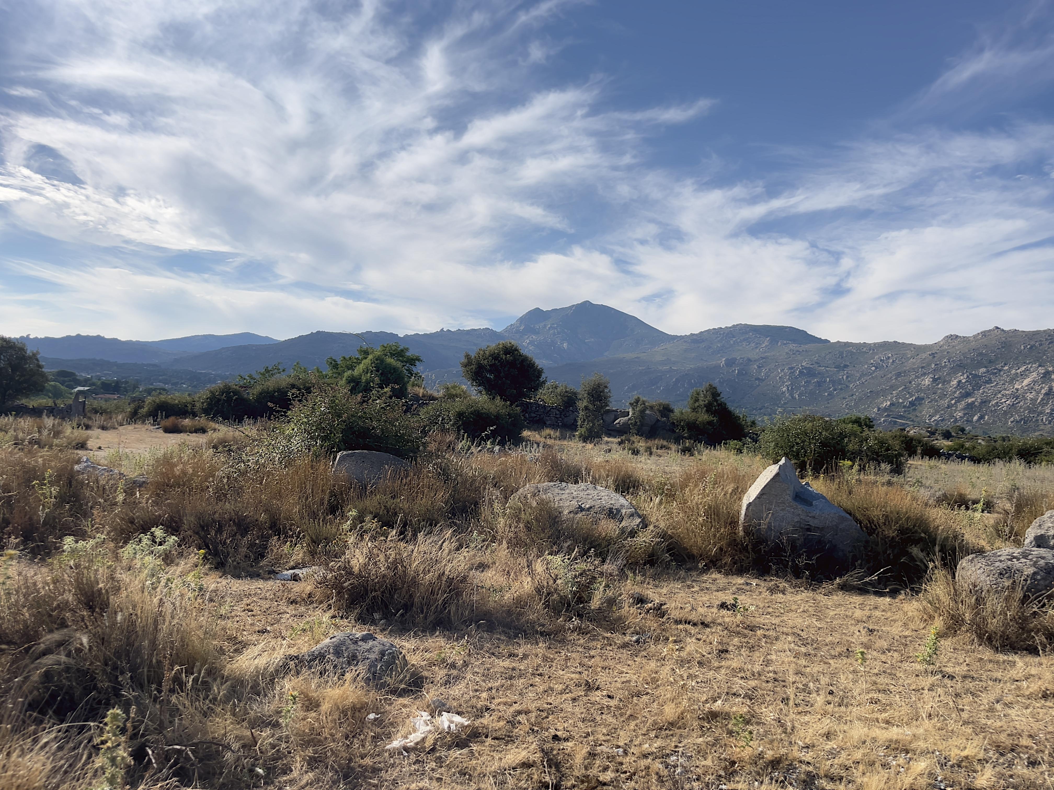 Dry, grassy field with scattered rocks and bushes in the foreground. Mountains rise in the background under a sky filled with wispy clouds.