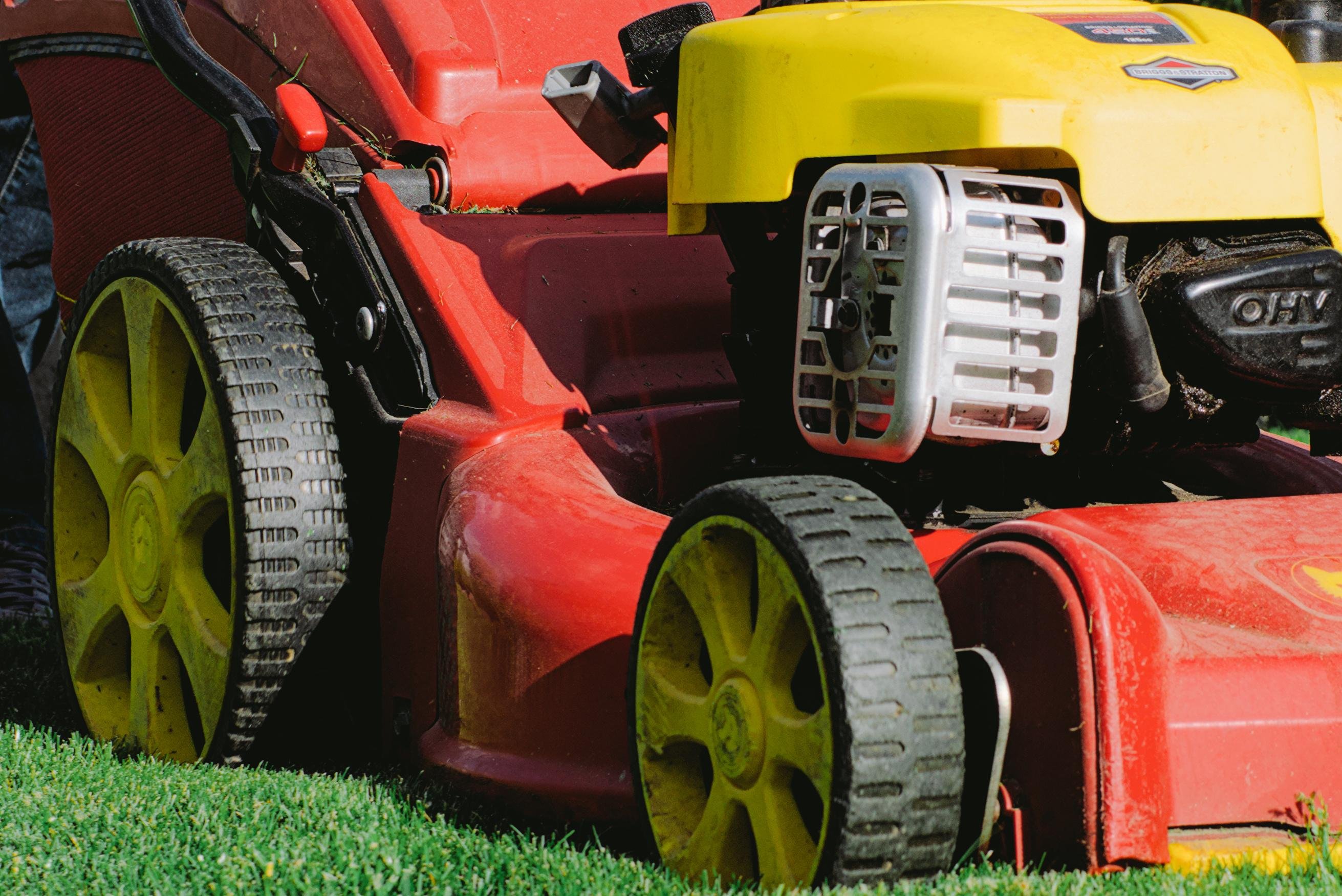 The image shows a close-up of a lawn mower. It features a red body with yellow accents, including the wheels and engine cover. It's positioned on grass, indicating it's likely being used for mowing.