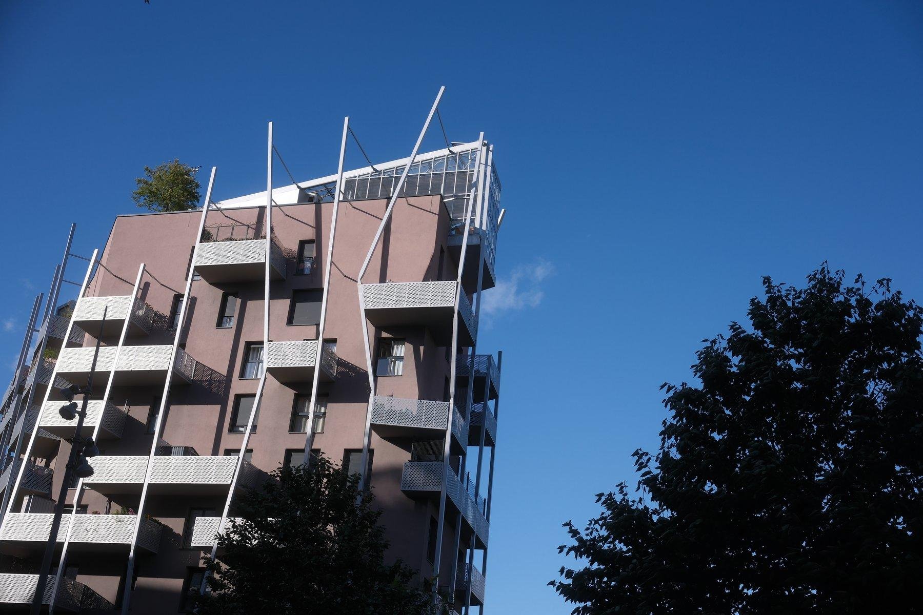 Photo horizontale. Un immeuble rose très moderne, avec des balcons avec rambardes en métal mais pas disposés de manière régulière, comme les fenêtres, et au sommet, une espèce de grande véranda en trapèze donnant à l’ensemble une silhouette non rectangulaire. Le ciel est bleu et le haut d’arbres sont visible en bas à droite de la photo