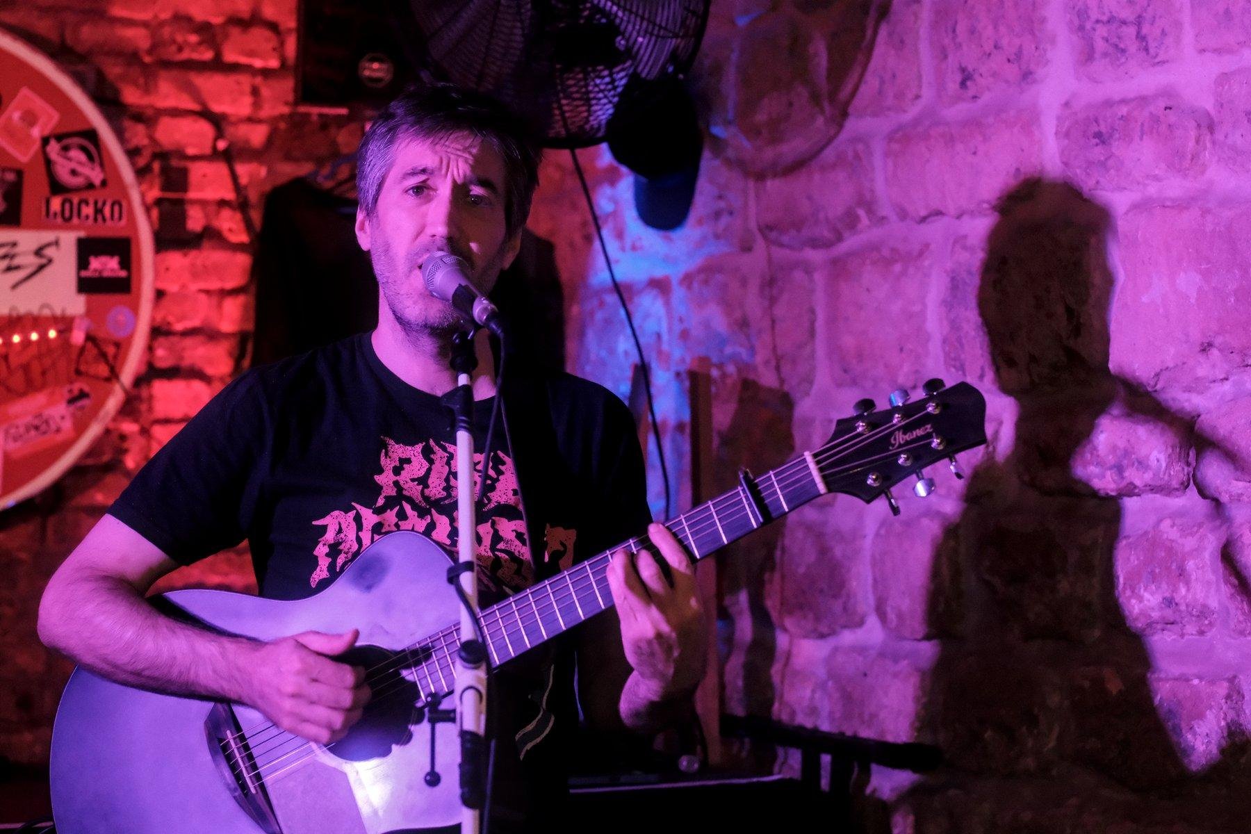 Photo horizontale. Portrait d’un homme aux cheveux cours, t-shirt noir avec des lettre rouges, jouant de la guitare en chantant. La lumières est rouge derrière lui et violette sur le mur de droite, où son ombre est projetée