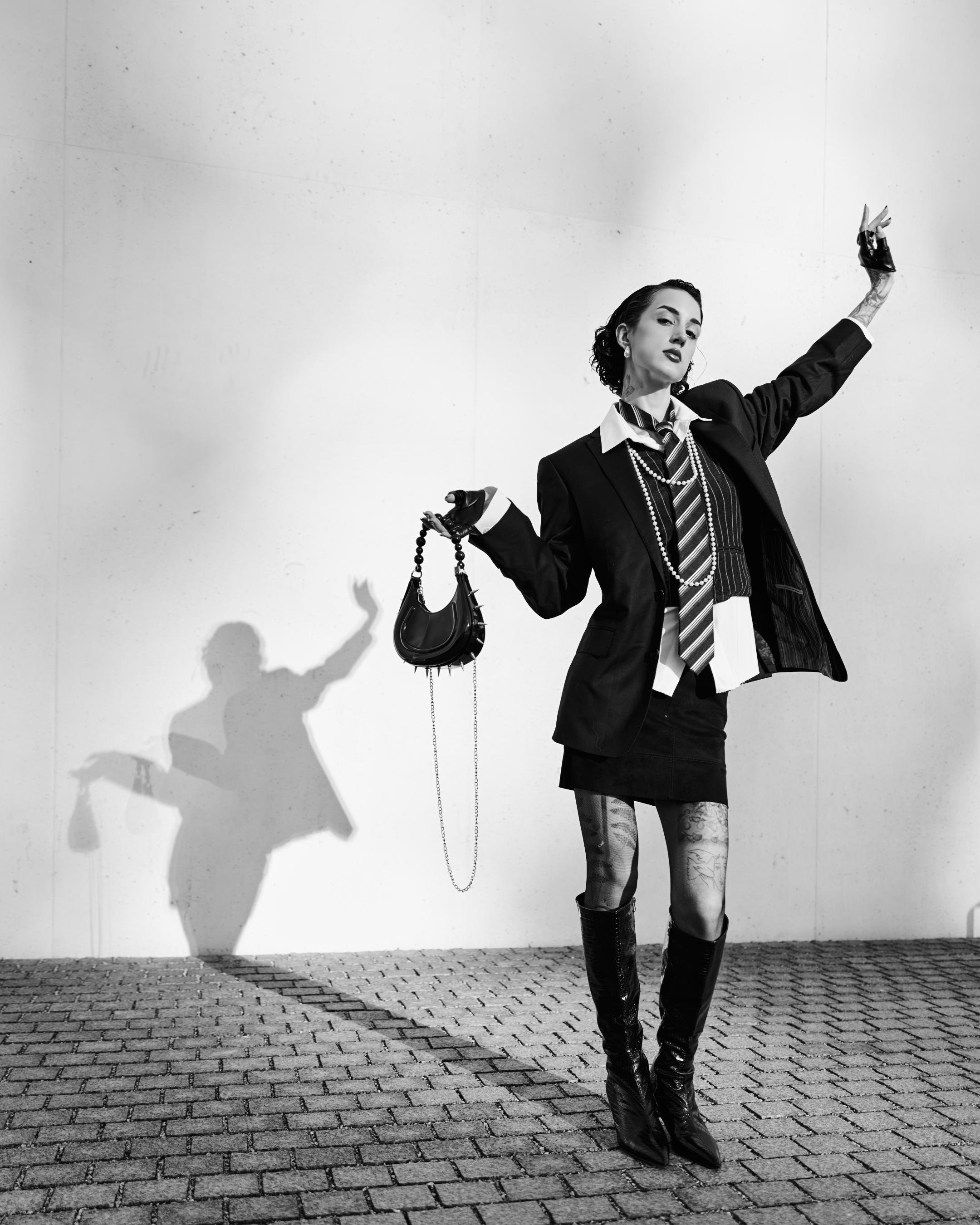 Black-and-white photo of a person posing on a brick-paved ground against a plain wall, wearing an oversized blazer, striped tie, short skirt, layered necklaces, and knee-high boots, holding a small handbag with a chain strap; their shadow is cast on the wall to the left.