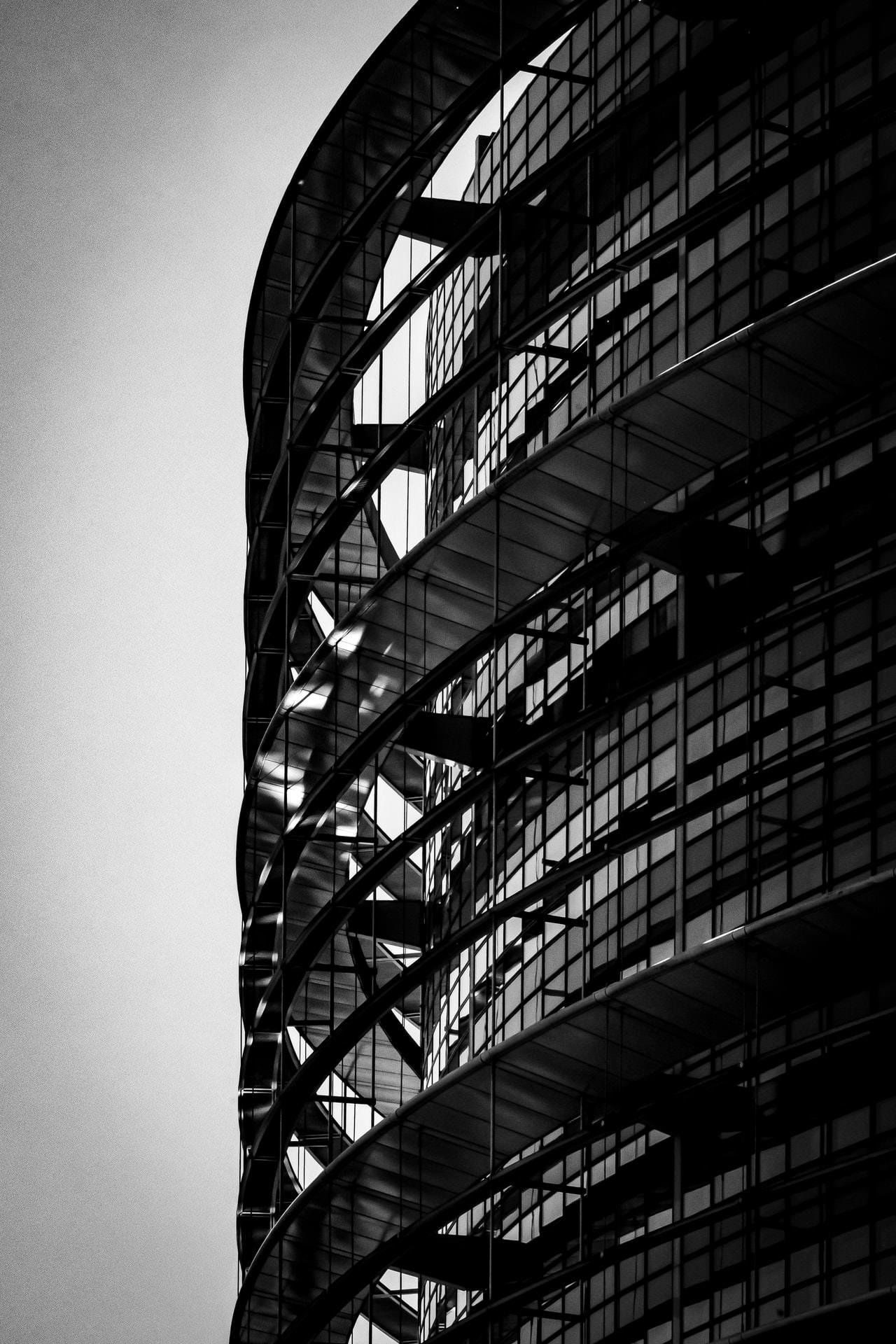 Curved facade of a modern building in black and white, featuring multiple floors with large glass windows and distinct horizontal lines. The structure includes reflective glass panels and metal framework, creating a repetitive geometric pattern. Bright backlighting highlights the contrast between the building and the sky.