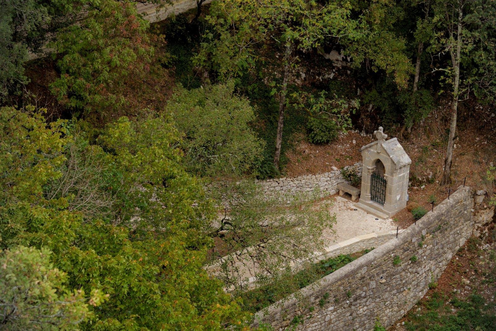 Photo horizontale. Un chemin en pierre qui mène à un petit mausolée surmonté d’une croix chrétienne. Un petit banc en pierre se trouve à gauche. Beaucoup d’arbres occupent l’image