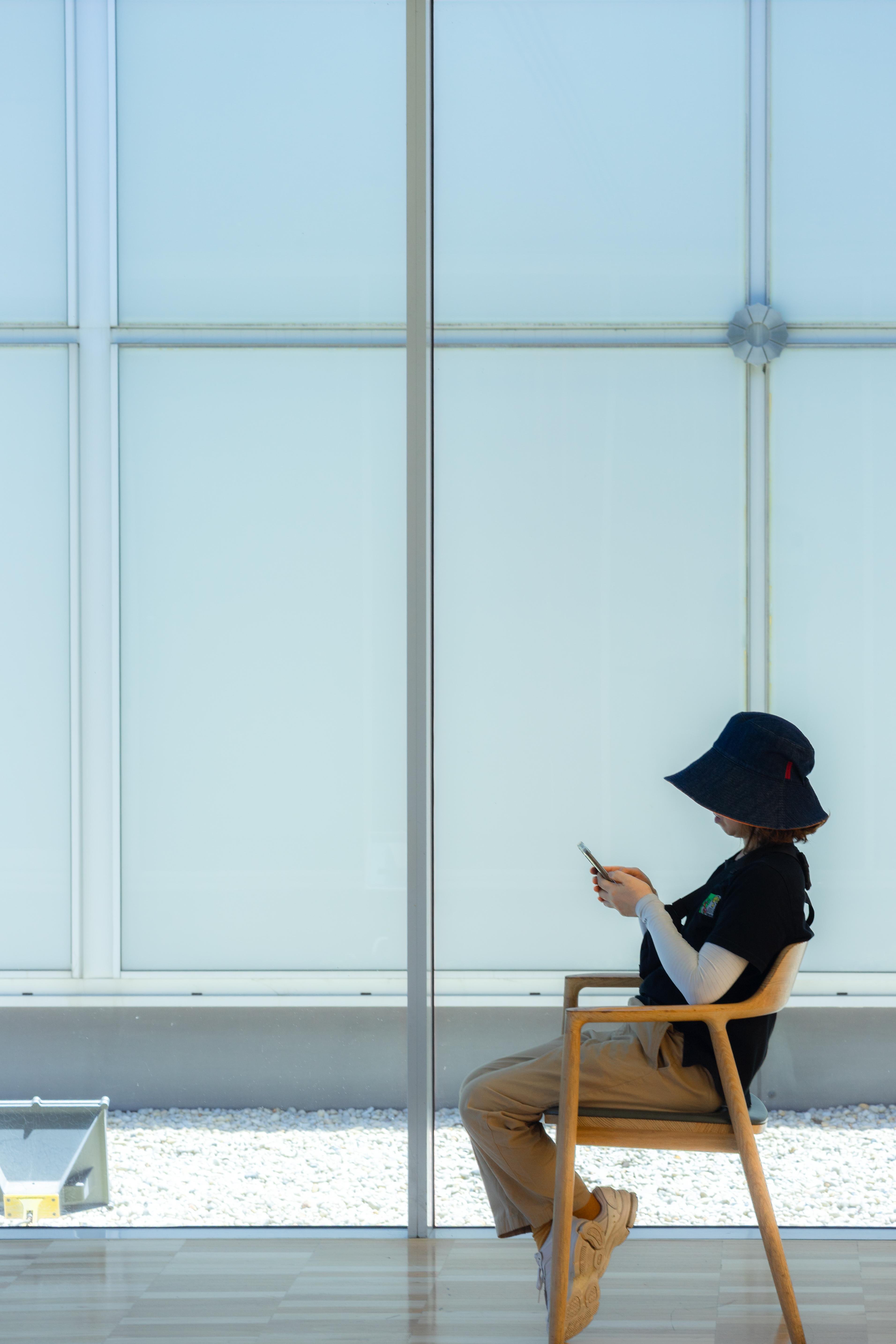 Seated person wearing a wide-brimmed hat, black shirt, and beige pants, looking at a phone. The setting includes large, frosted glass windows and a wooden floor. The scene is minimalistic with natural lighting.