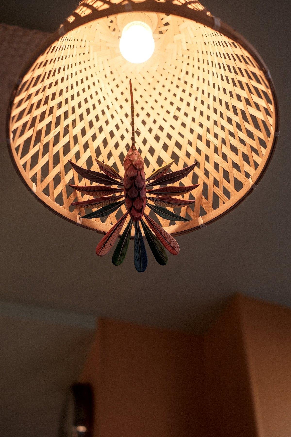 Photo verticale. Contre plongée sur un plafonnier dont l’abat jour est un cone en quadrillage de bois. Sur le cercle, un colibri en bois est accroché, donnant l’impression qu’il butine la lampe