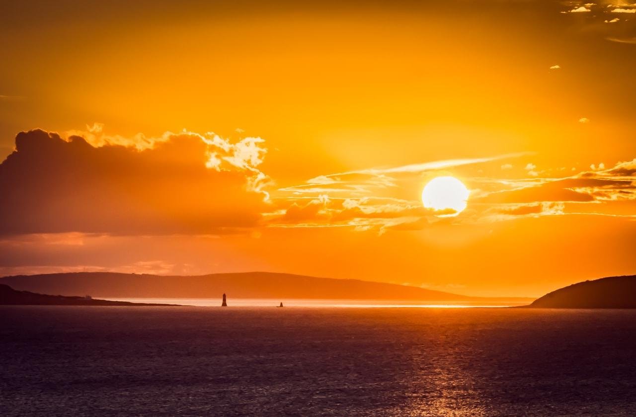 This image depicts a scenic sunset over the ocean. The sky is illuminated with warm orange and golden hues, and the sun is partially visible above the horizon. Clouds are scattered, adding texture to the sky. The silhouette of distant land and possibly a lighthouse or sailboat can be seen on the horizon. The ocean surface reflects the colors of the sunset, creating a serene and picturesque view.