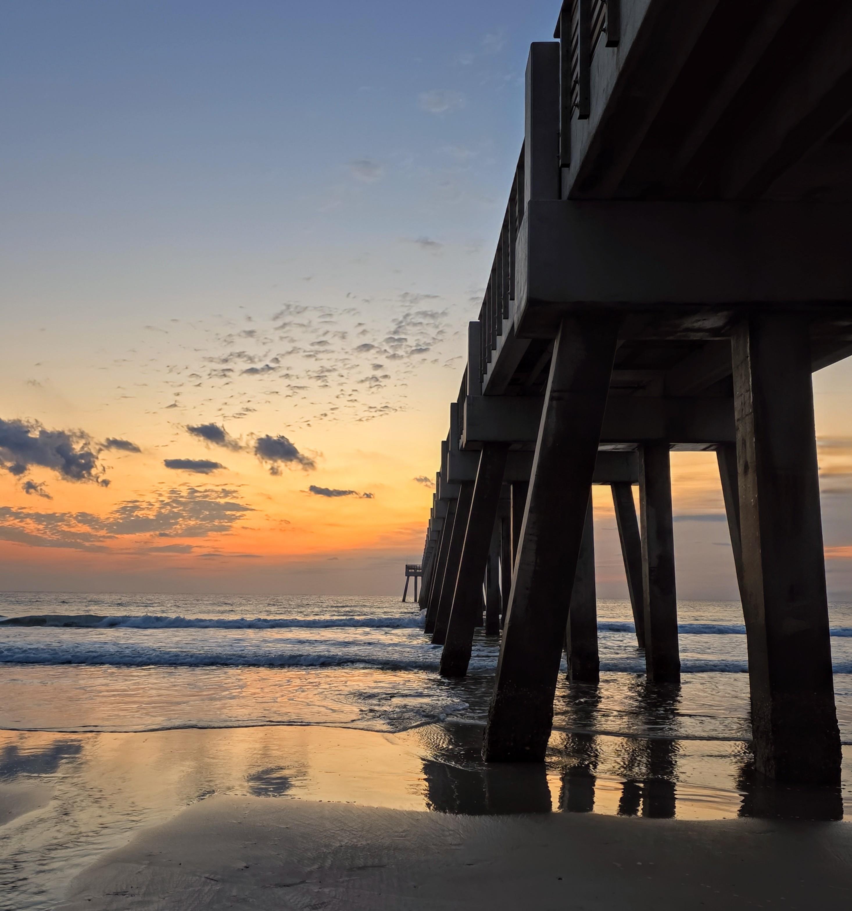 Sunrise over the ocean with a long pier extending from the right side. The sky displays shades of orange and blue, scattered with small clouds. Waves gently lap against the sandy shore, reflecting the pier and colorful sky on the wet sand.
