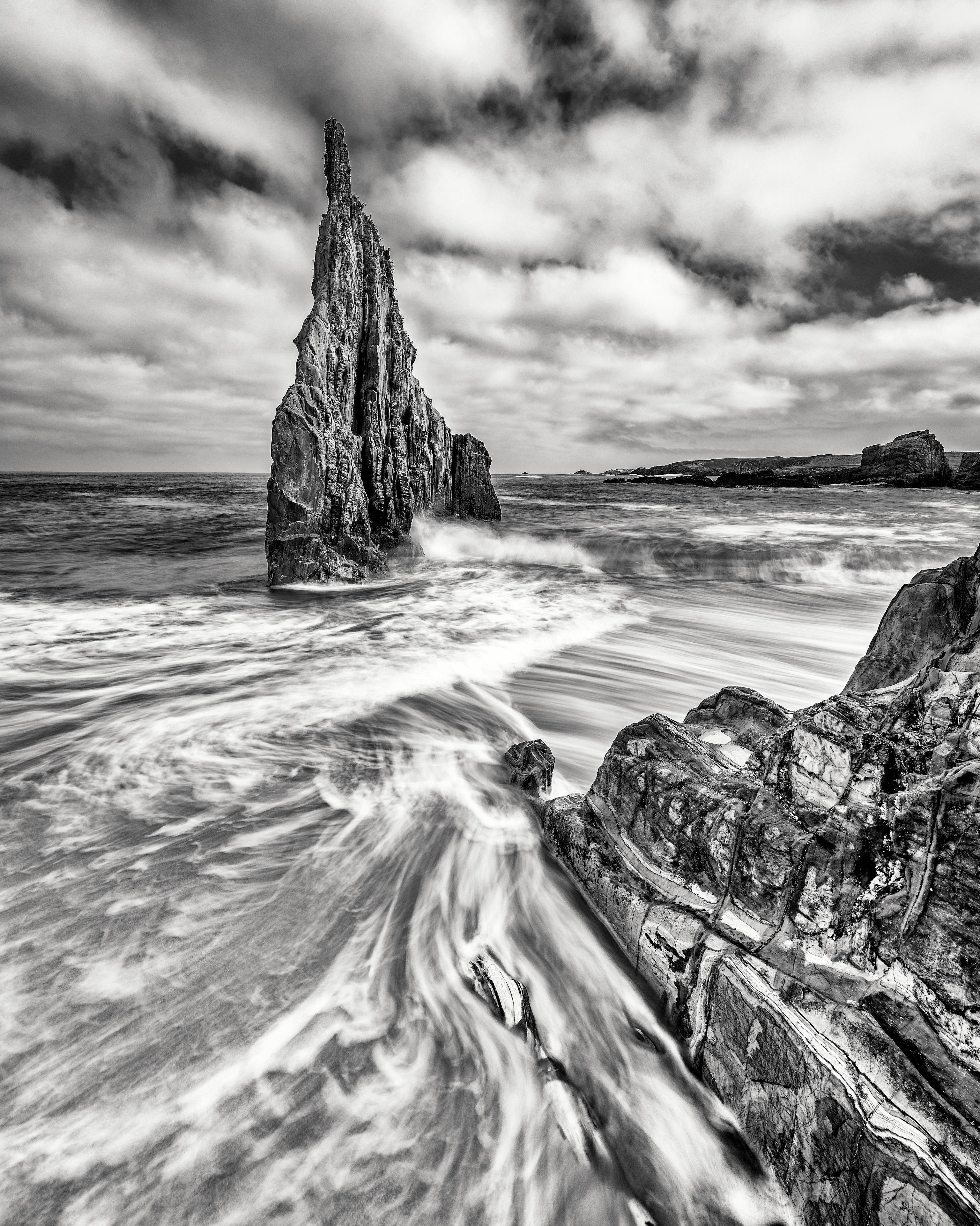 Dramatic black and white coastal scene with a towering, jagged rock formation rising from the sea. Waves swirl dynamically around the base, creating streaks of white foam. The foreground features textured rocks, and the sky is filled with scattered clouds, adding depth to the image.