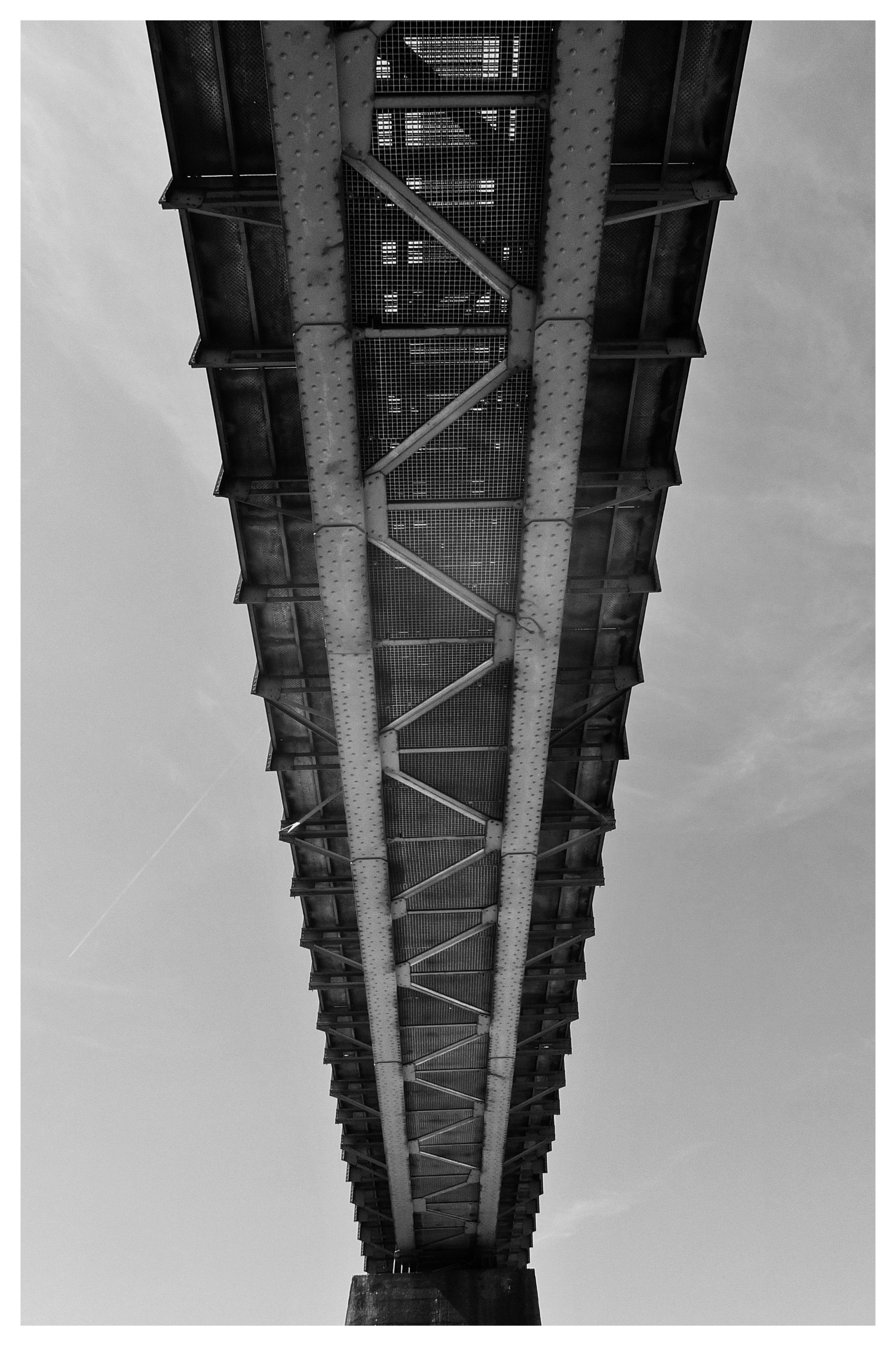 Underside view of a steel bridge centered in the frame, showing riveted beams, cross-bracing, and a grated walkway, narrowing toward a concrete support against a cloudy sky.