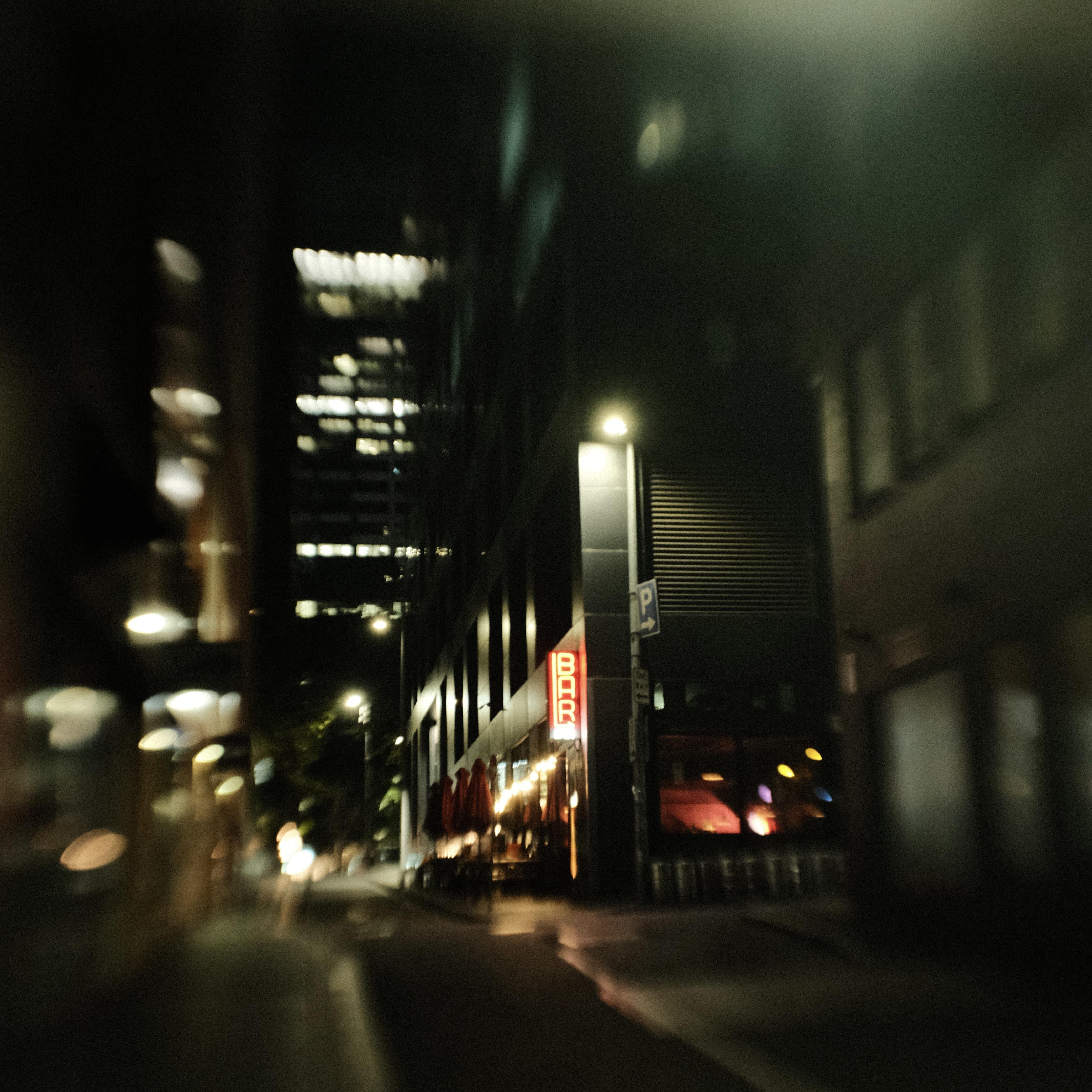 Blurred nighttime city street with tall buildings; a red vertical neon “BAR” sign glows on a corner storefront under streetlights, with scattered window lights and indistinct traffic in the distance.