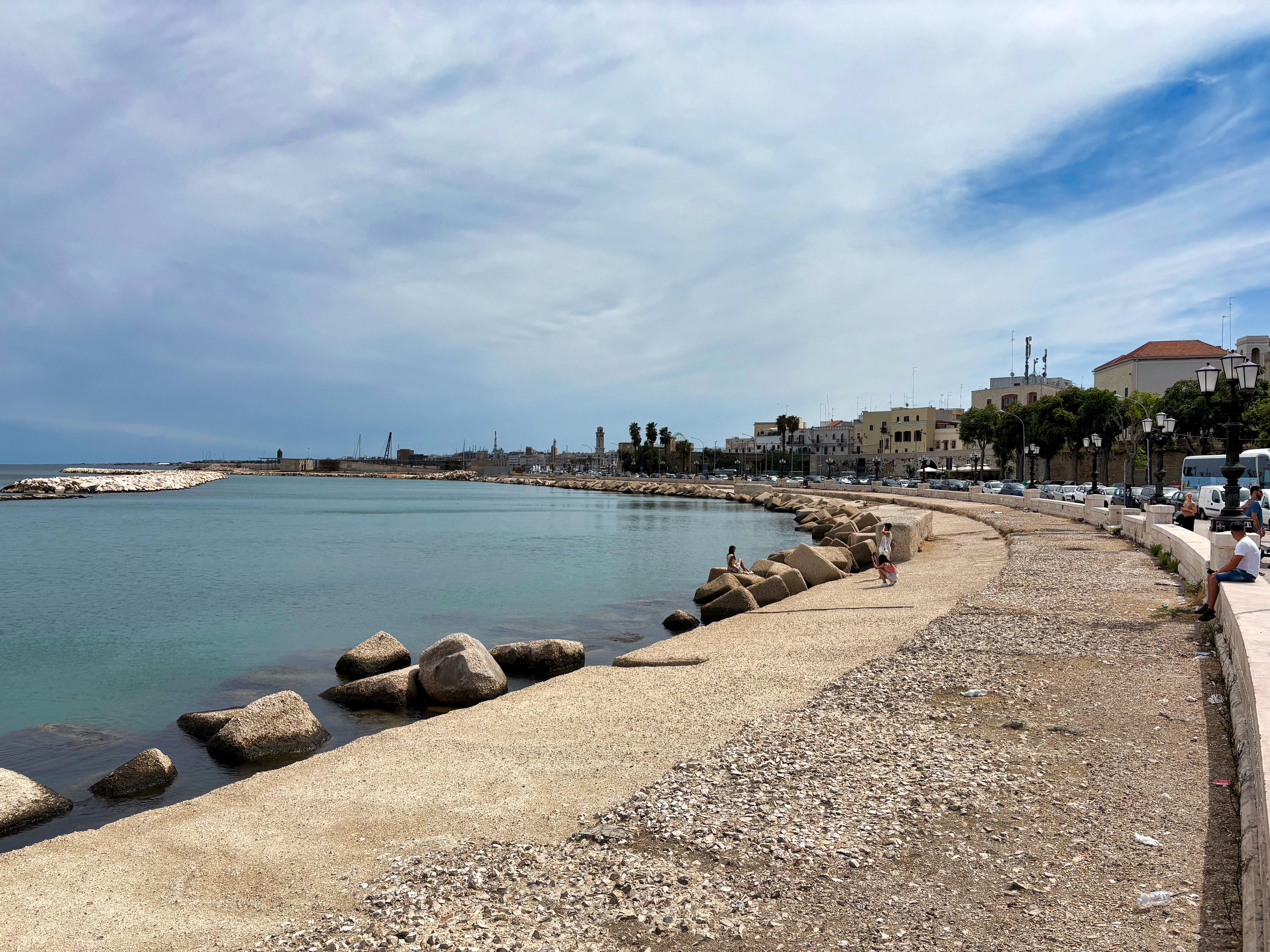 Coastal promenade with a wide walkway and large rocks lining the shore. Calm blue waters stretch into the distance with buildings visible along the waterfront. People are seated and walking along the promenade. The sky is partly cloudy with patches of blue.