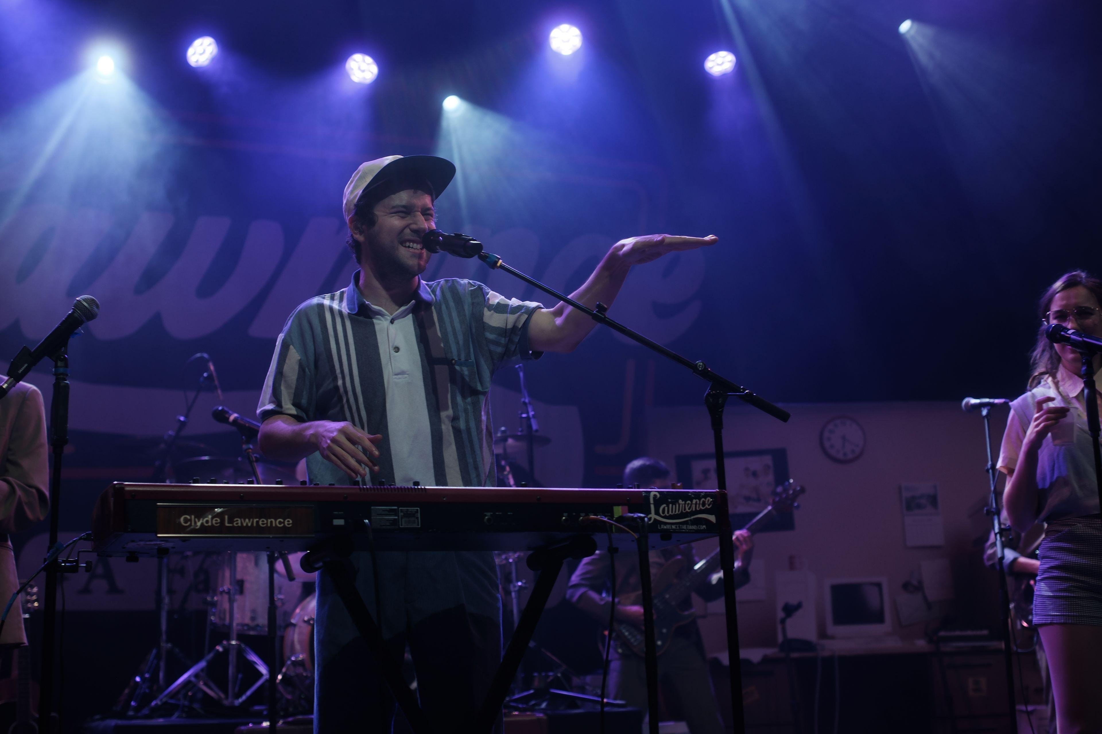 Photo horizontale. Clyde Lawrence sur scène, derrière son clavier électronique, en train de mimer la hauteur d’une note avec sa main gauche à plat dans les airs